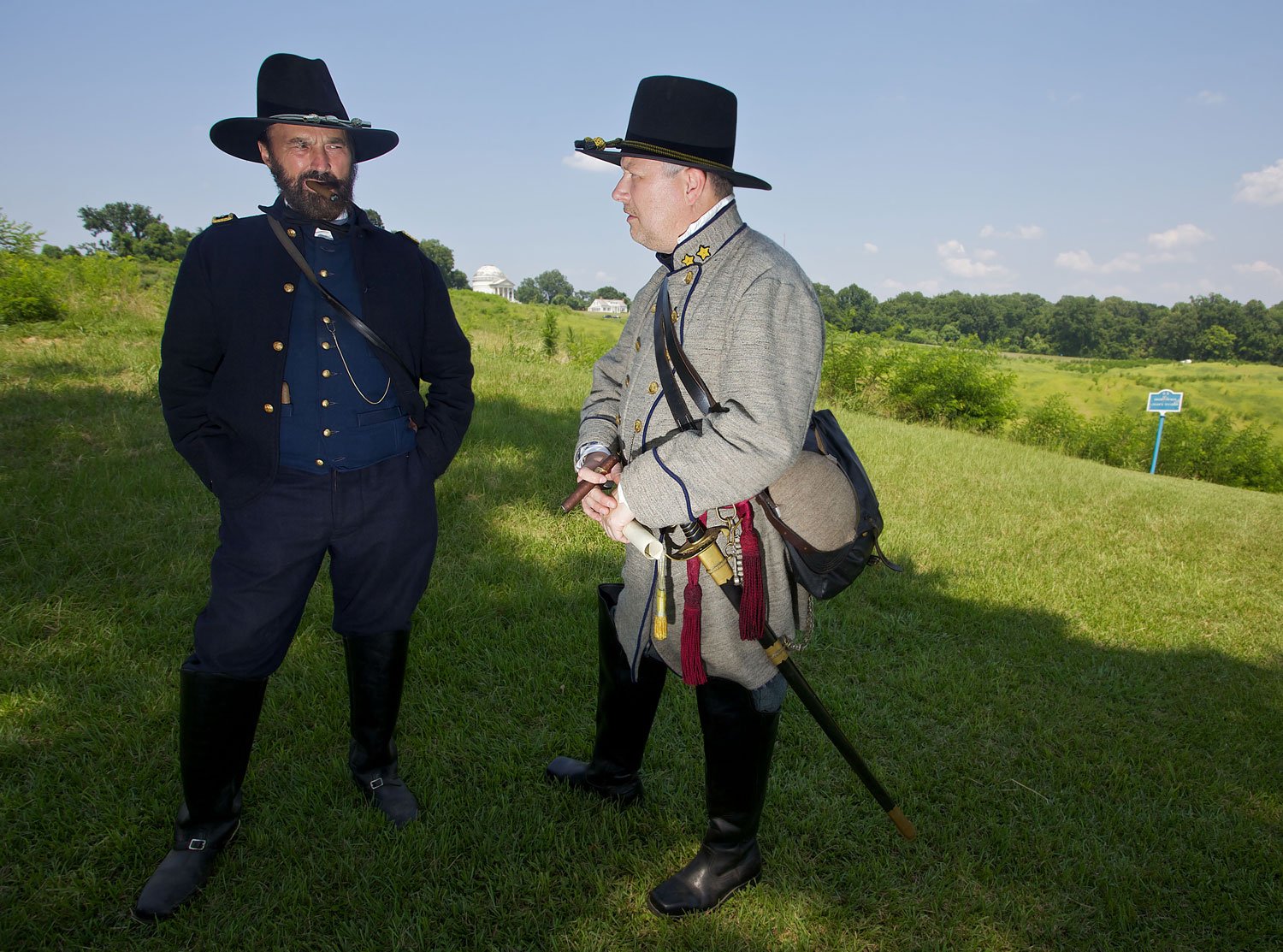 Two men dressed as Civil War soldiers standing outdoors on a grassy field, engaged in conversation under a bright blue sky with trees and buildings in the background.