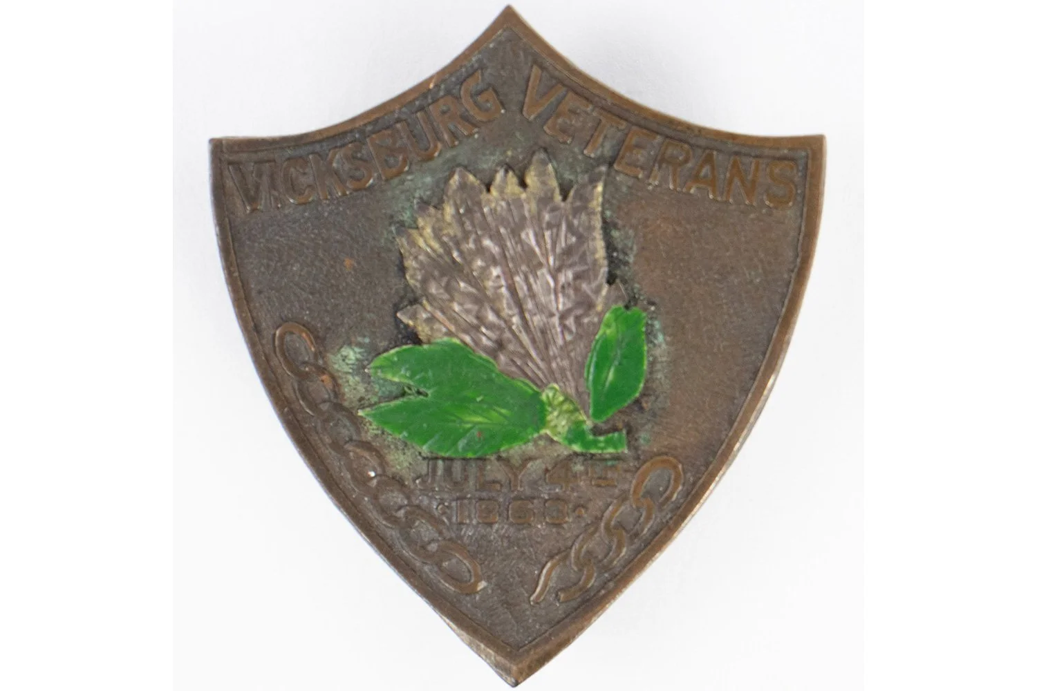 A vintage metal badge with a shield shape, featuring a painted brown flower with green leaves in the center and the text "Vicksburg Veterans" at the top.