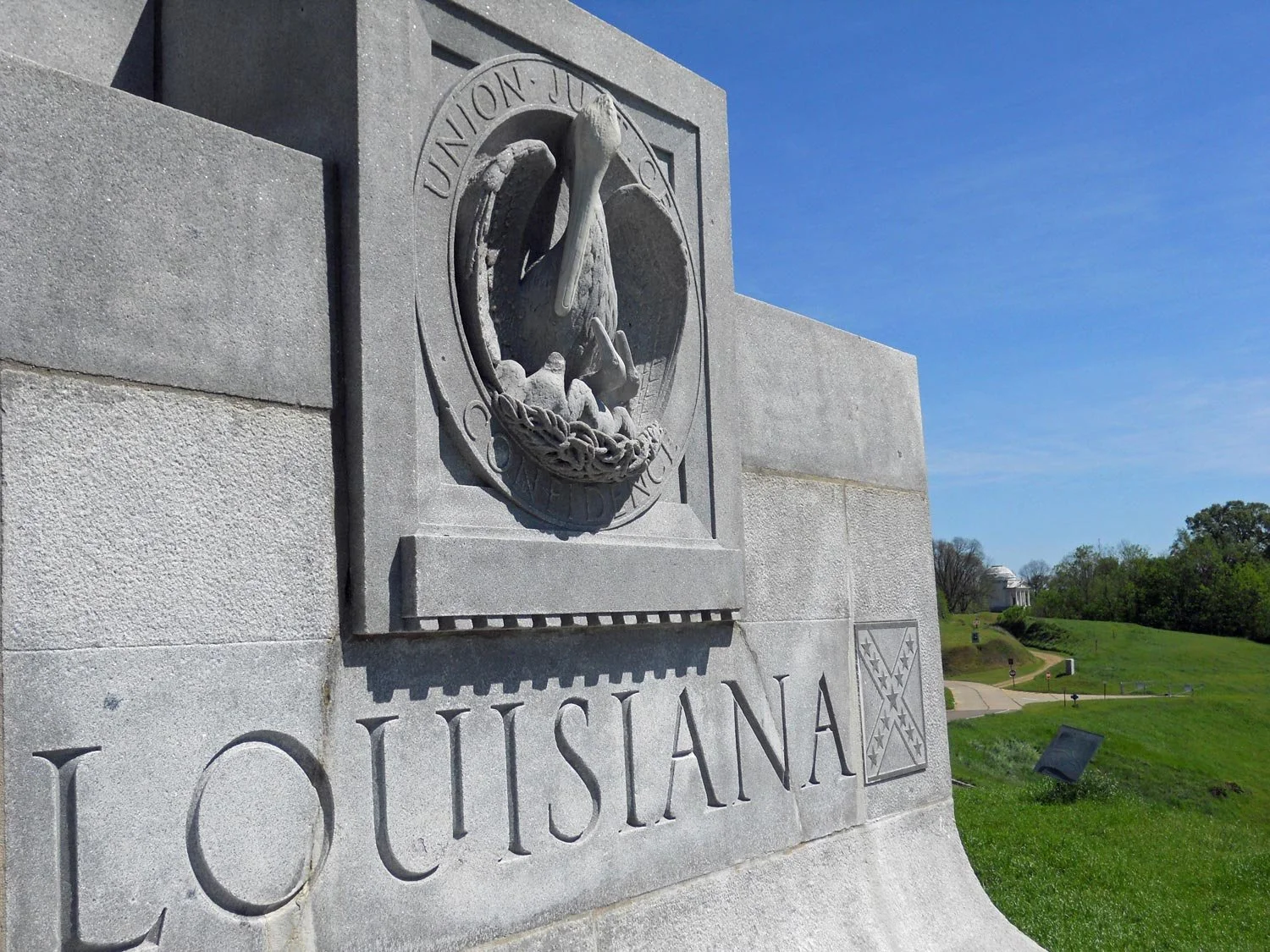 The image shows a stone monument with a carved emblem of a pelican feeding her chicks and the word 'LOUISIANA' inscribed below. In the background is a green landscape with trees and a white building with a dome.
