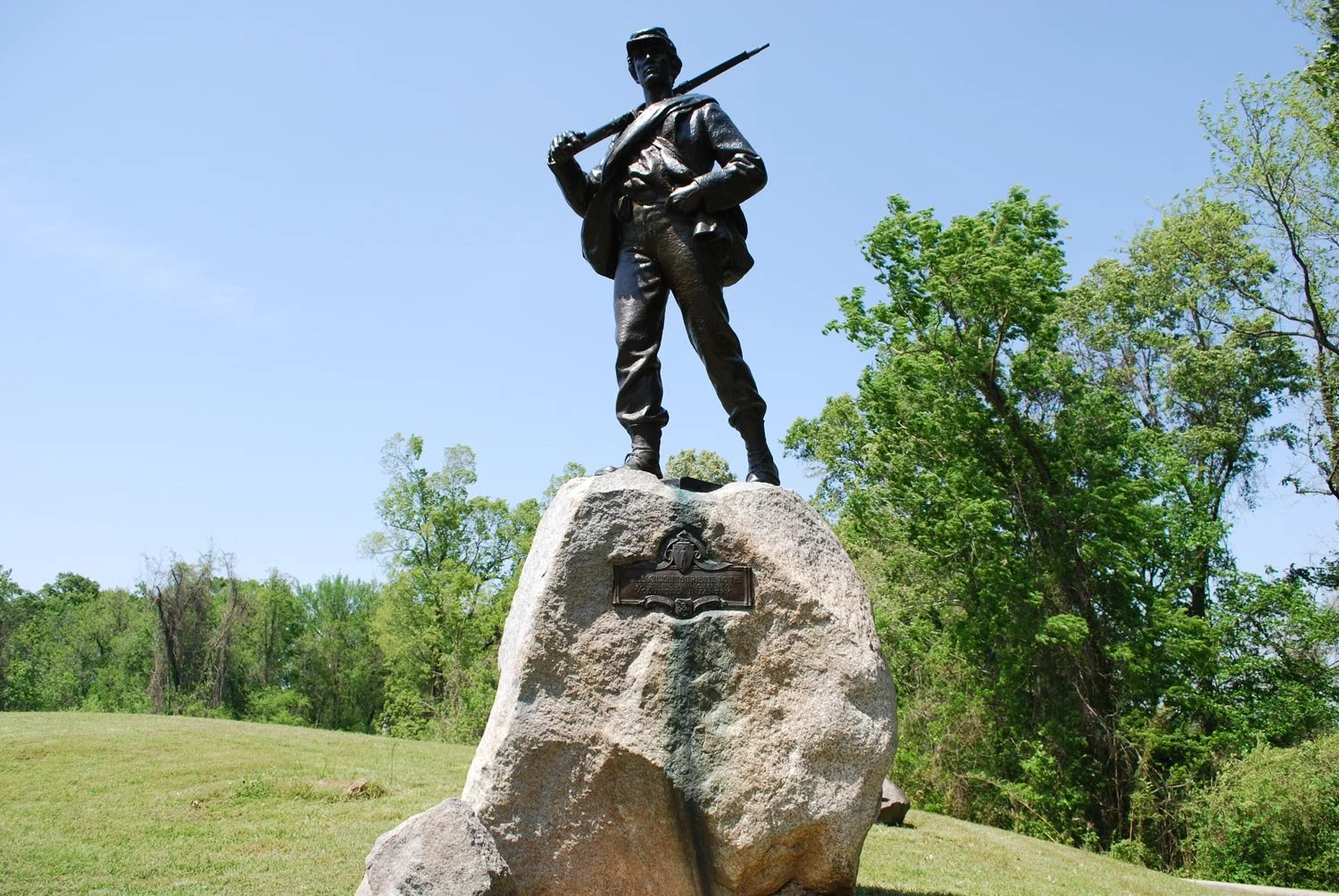 Statue of a soldier in military uniform standing on a large rock, holding a rifle over his shoulder, with trees and a clear blue sky in the background.