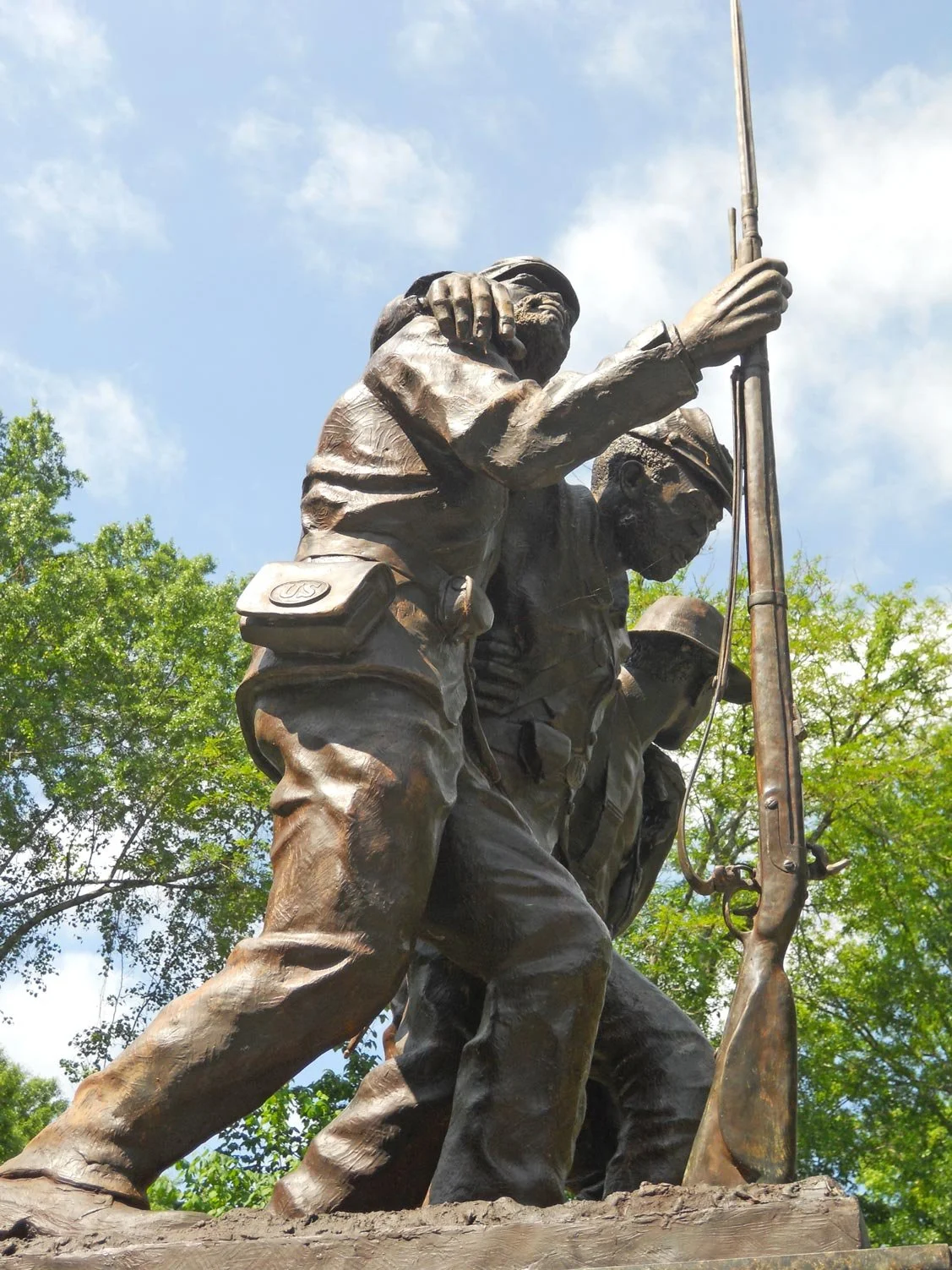 Bronze statue of two soldiers, one carrying a rifle, set outdoors with trees and blue sky in the background.