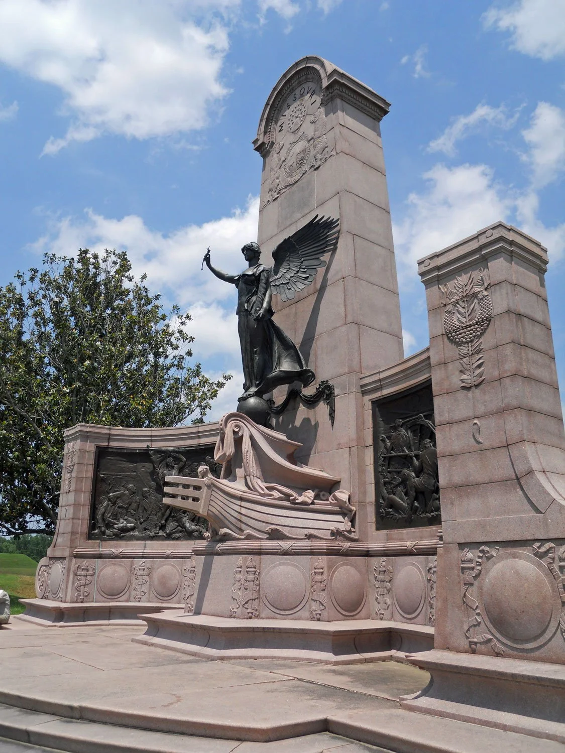A monument featuring a winged female figure holding a torch, with detailed carvings and a tall stone column against a partly cloudy sky.