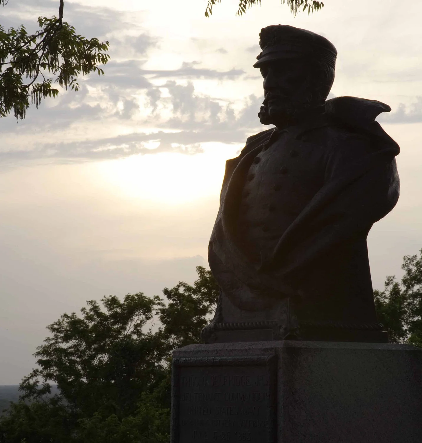 Silhouette of a bronze statue of a man wearing a military uniform and cap, with trees and a cloudy sky in the background.