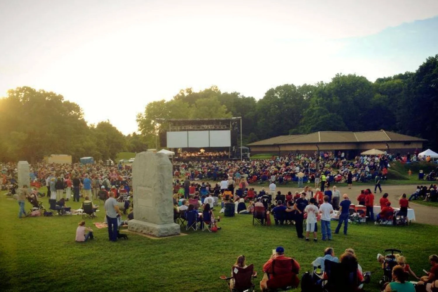Large outdoor gathering at a park with a stage, many people sitting on the grass, some in chairs, watching a movie or presentation on a large screen, surrounded by trees, during sunset.