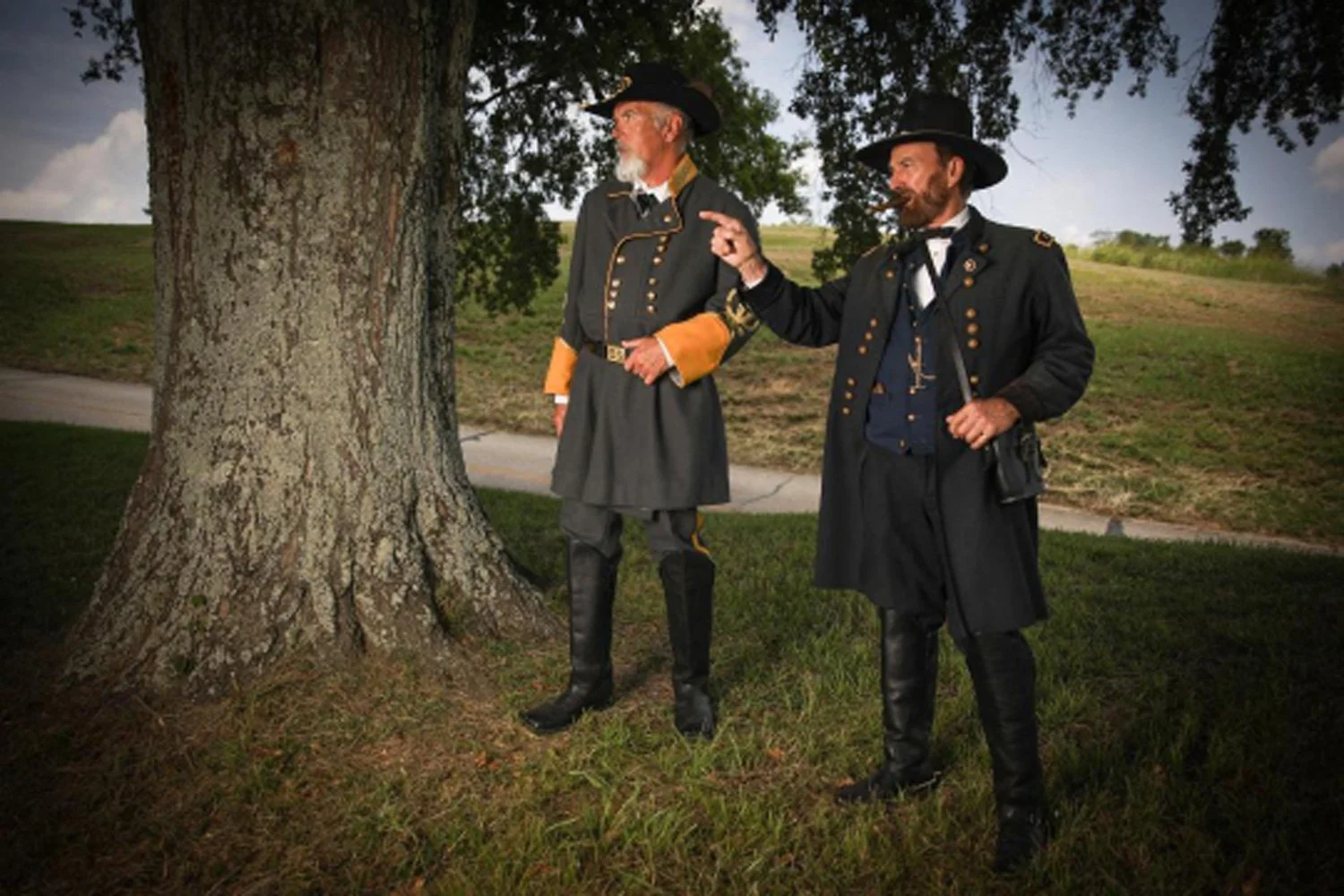 Two men dressed as soldiers or officers from historical period, wearing black coats, tall hats, and leather boots, standing outdoors near a large tree. One man is pointing, and smokes a cigar.