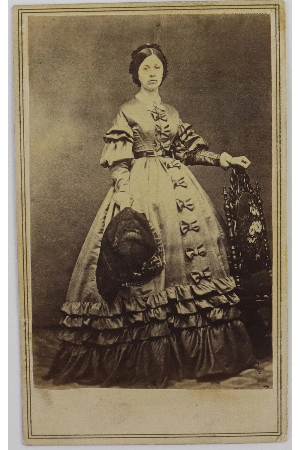A historical sepia photograph of a woman from the 19th century wearing a Victorian-style dress with bows and ruffles, standing next to a small ornate chair.