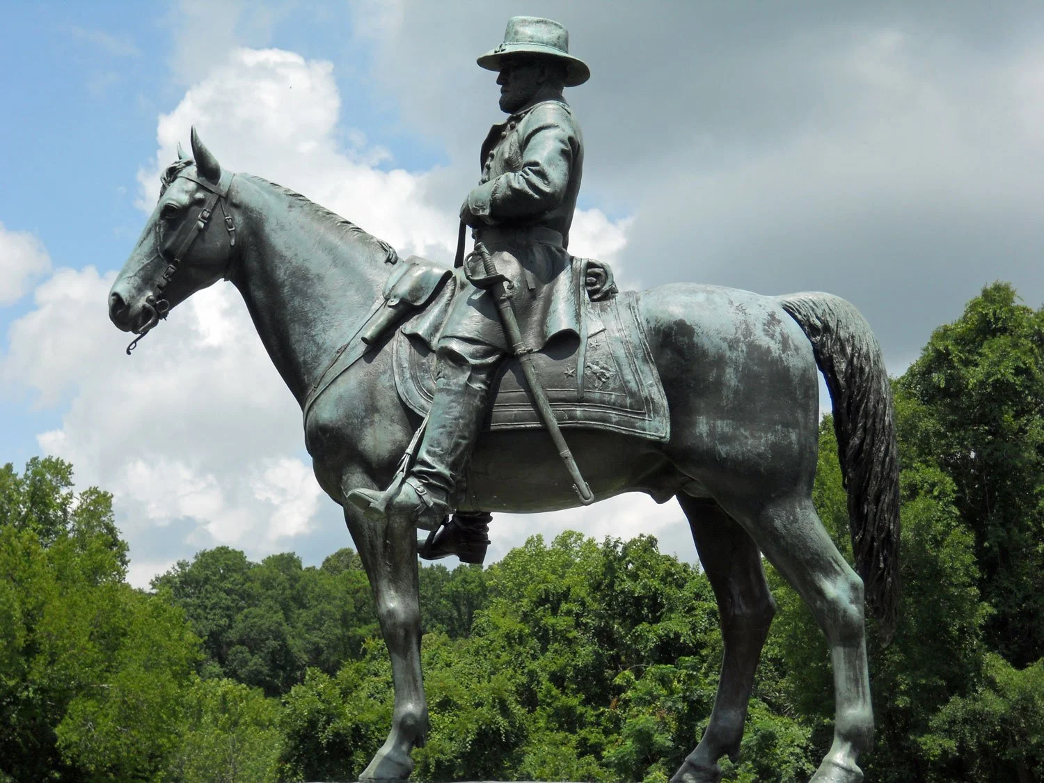 Bronze statue of a man in a military uniform riding a horse outdoors with trees and clouds in the background.