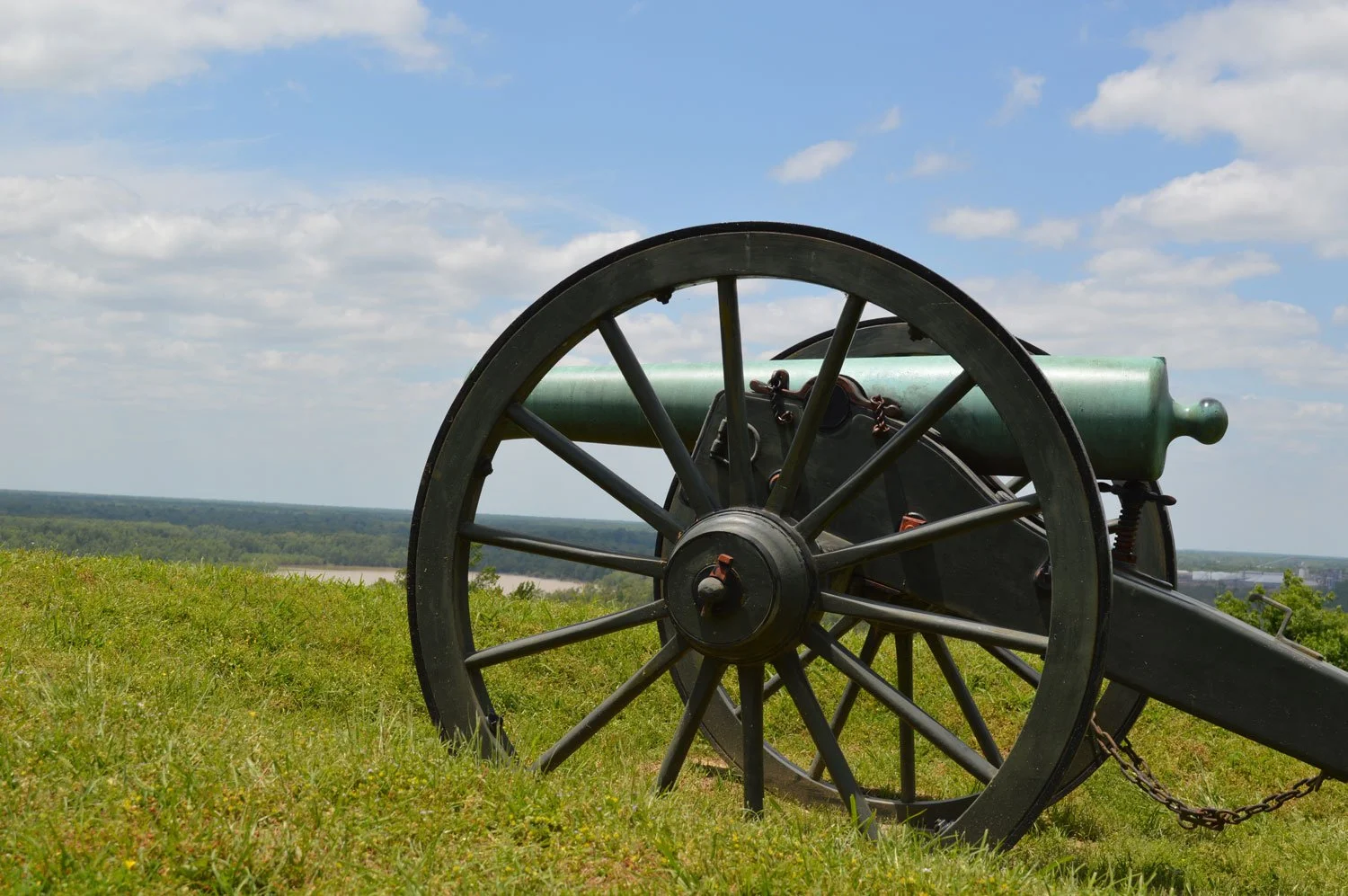 An old green cannon with black wheels sitting on a grassy field under a partly cloudy sky.