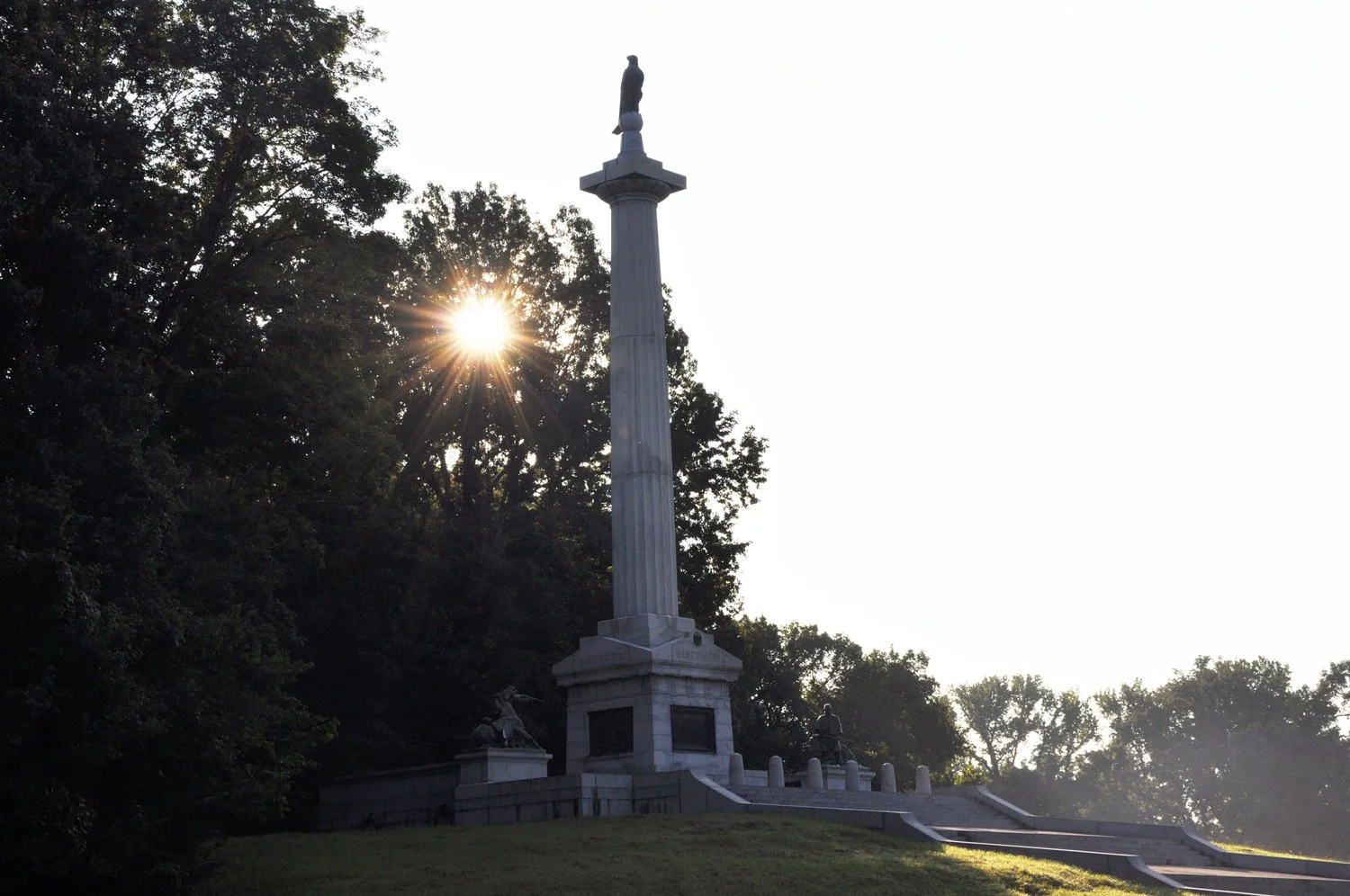 A monument with a tall column topped by a sculpture, surrounded by trees with sunlight peeking through them.