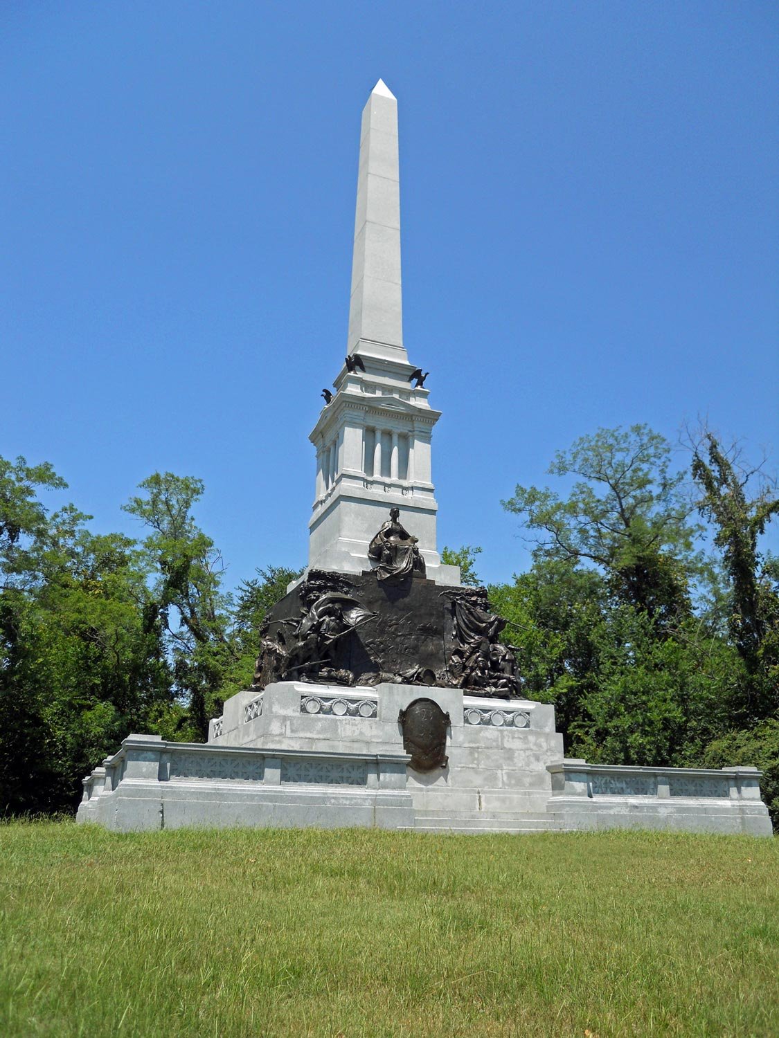 White monument with an obelisk on top, adorned with bronze sculptures and a seated bronze figure at the base, surrounded by green trees under a clear blue sky.
