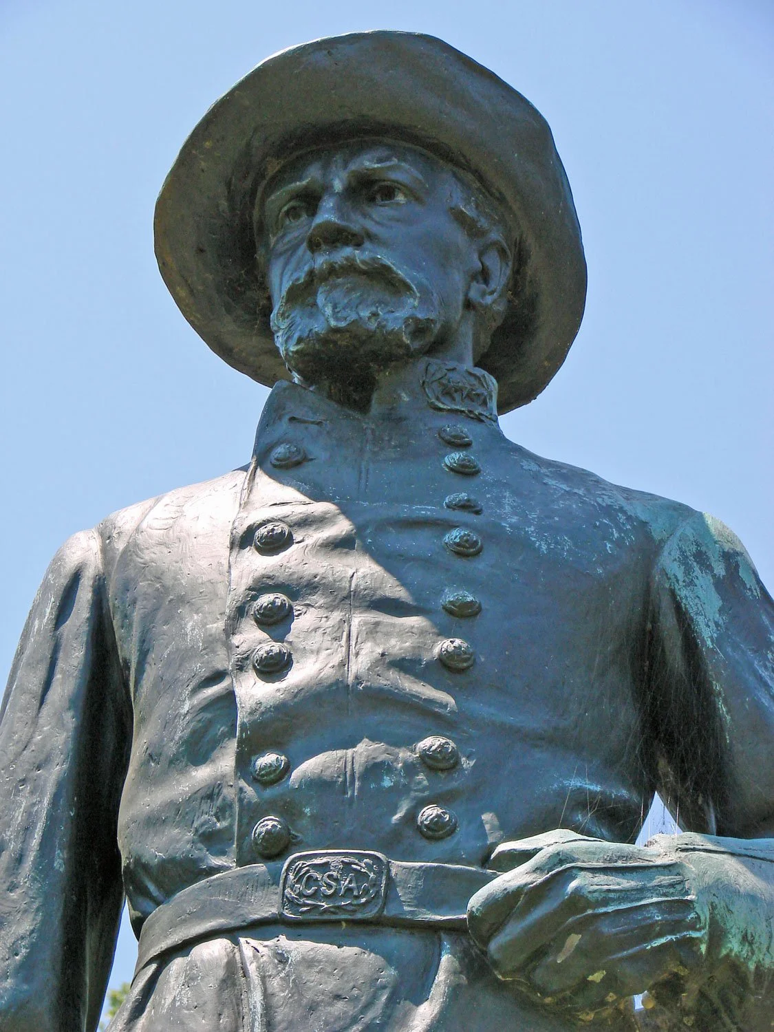 Close-up of a bronze statue of a man dressed in historical military attire, wearing a wide-brimmed hat, with detailed buttons on his coat and a sash with the letters 'CSA' at his waist, against a clear blue sky.