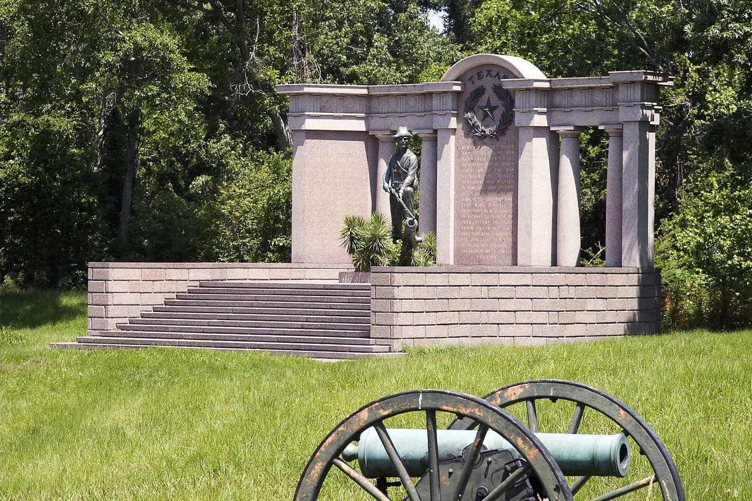 A war memorial monument featuring a statue of a soldier with a rifle, carved into a gray stone structure with columns and a plaque, surrounded by greenery, and a cannon with large wheels in the foreground.