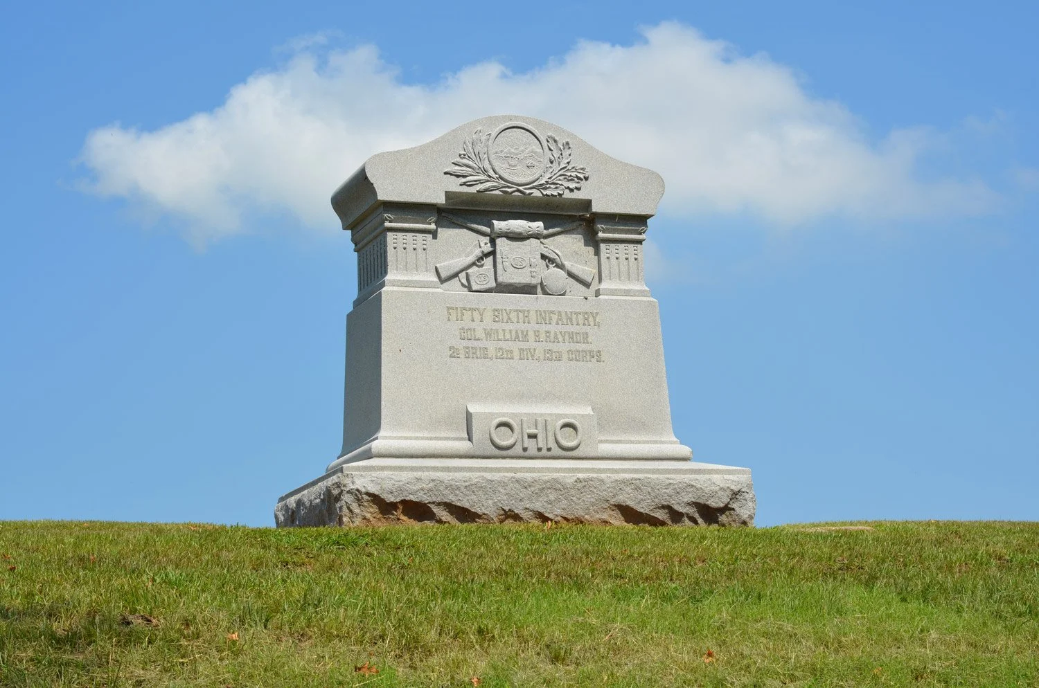 A historical monument dedicated to the Fifty Sixth Infantry, Colonel William M. Raynor in Ohio, featuring a granite pedestal with military symbols, set on a grassy hilltop against a blue sky with clouds.