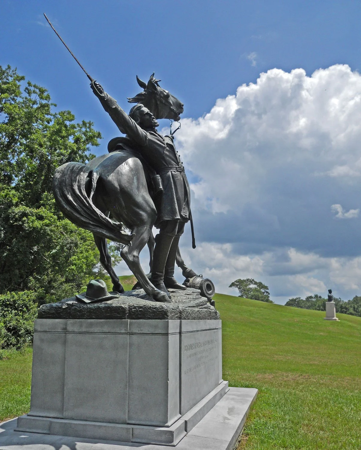 A bronze statue of a soldier on horseback, raising a sword skyward, with a hat placed on the pedestal in front. The scene is outdoors with green grass, trees, and a partly cloudy sky.