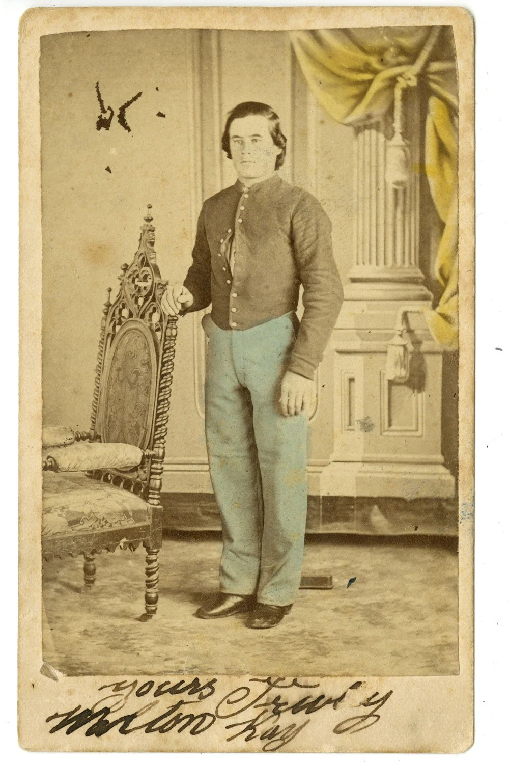 A black and white photo of a young man in Civil War-era clothing standing beside an ornate chair with a high back, in front of a backdrop with a draped curtain and pillar. The photo appears aged with some damage and handwritten text at the bottom.