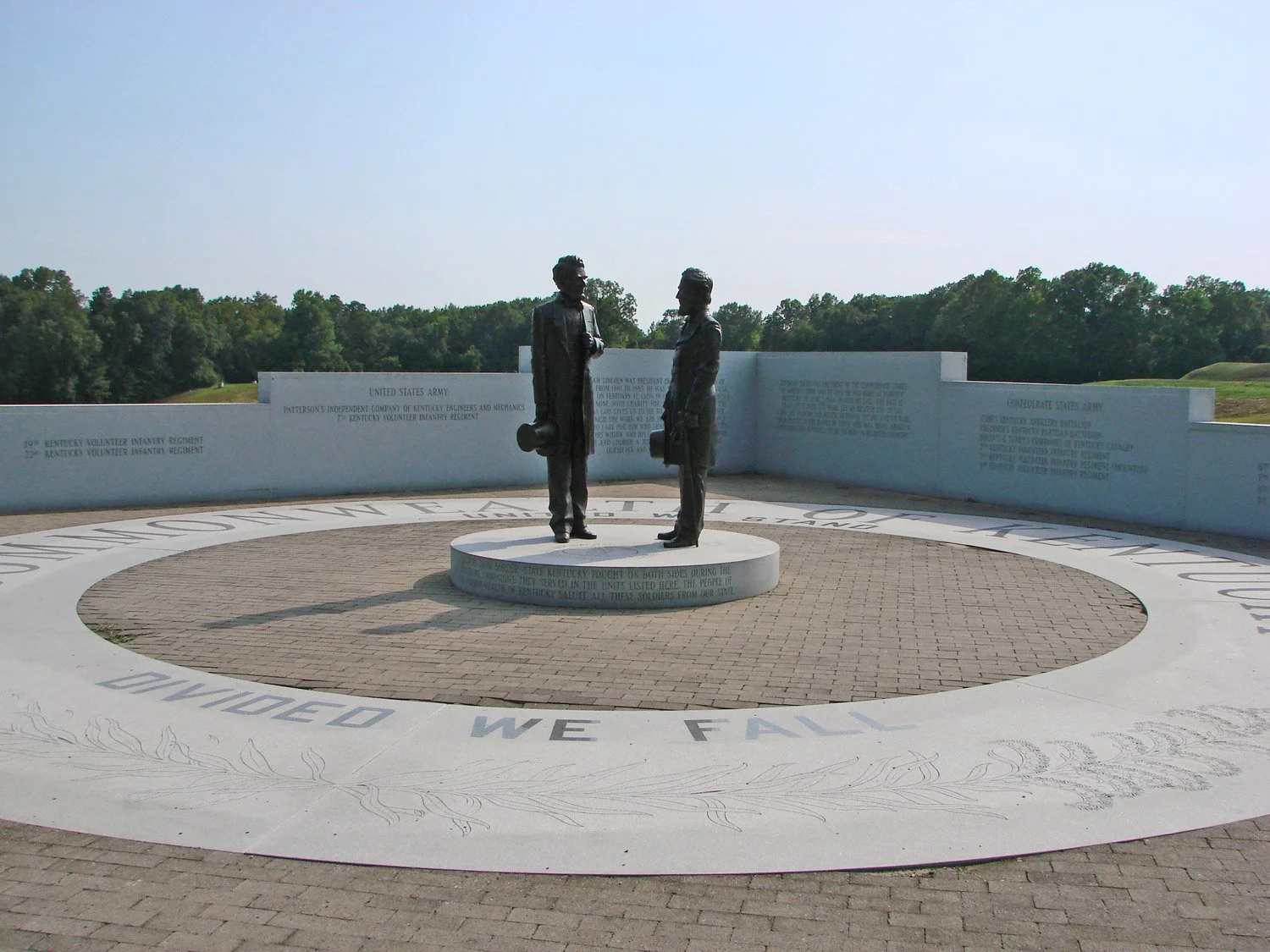 Statue of two soldiers shaking hands in a memorial with engraved text and a circular inscription on the ground, with trees in the background.