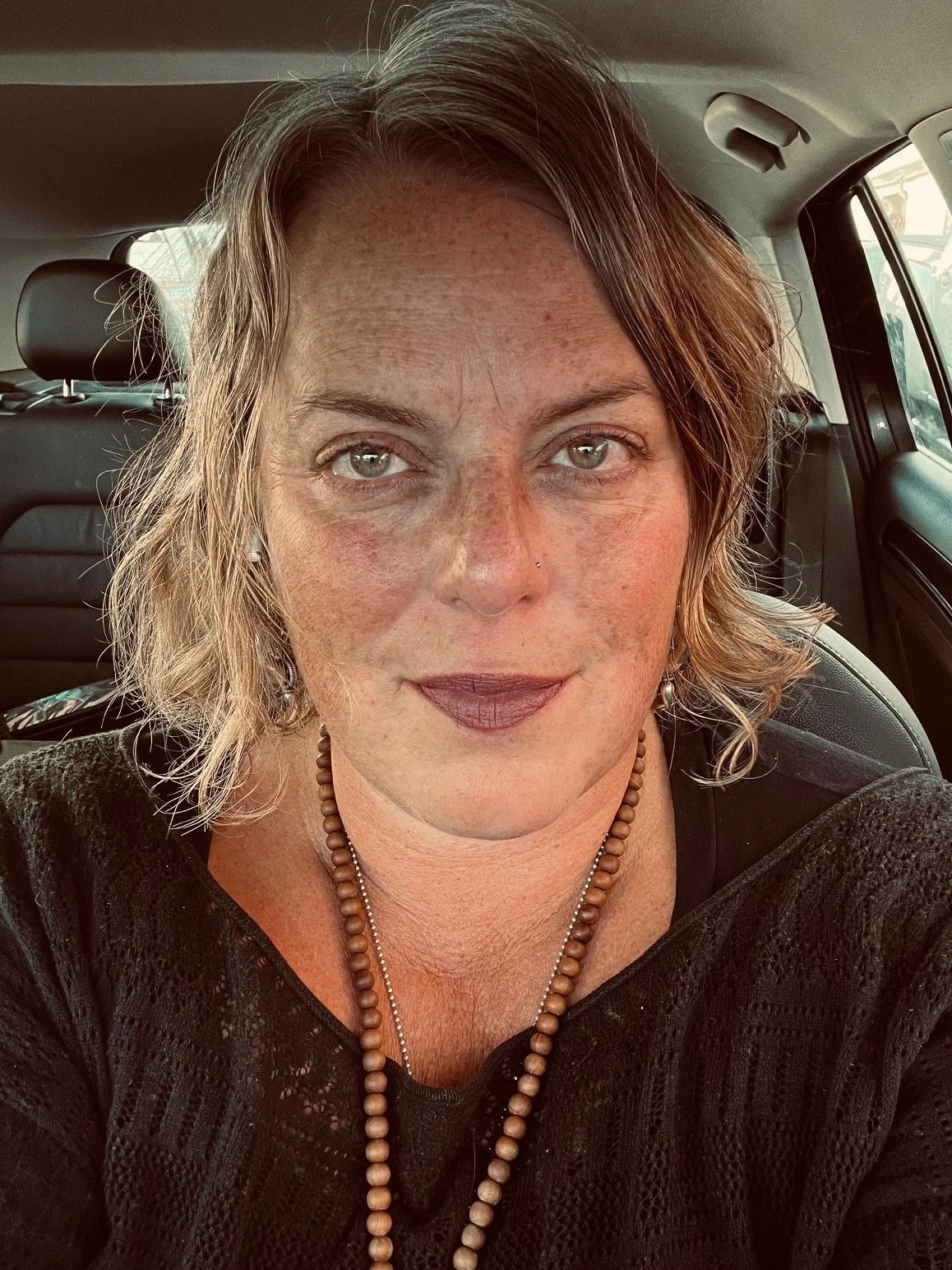 A woman with short, wavy hair and light-colored eyes, wearing dark lipstick, a black top, and beaded necklaces, sitting inside a vehicle.
