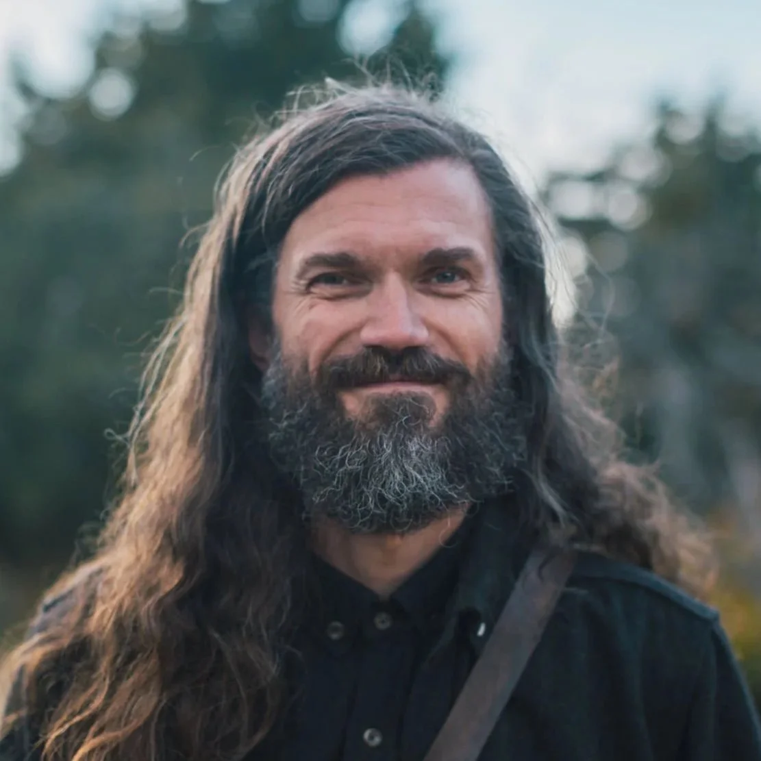 A man with long, dark hair and a beard smiling outdoors with trees in the background.