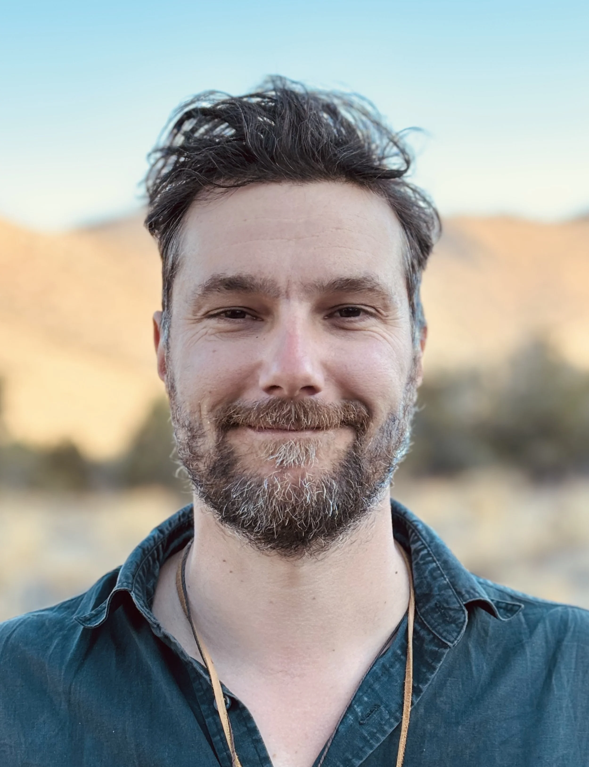 A man with short dark hair, a beard, and mustache smiling outdoors during daytime, wearing a dark green shirt and a necklace, with a blurred natural landscape in the background.