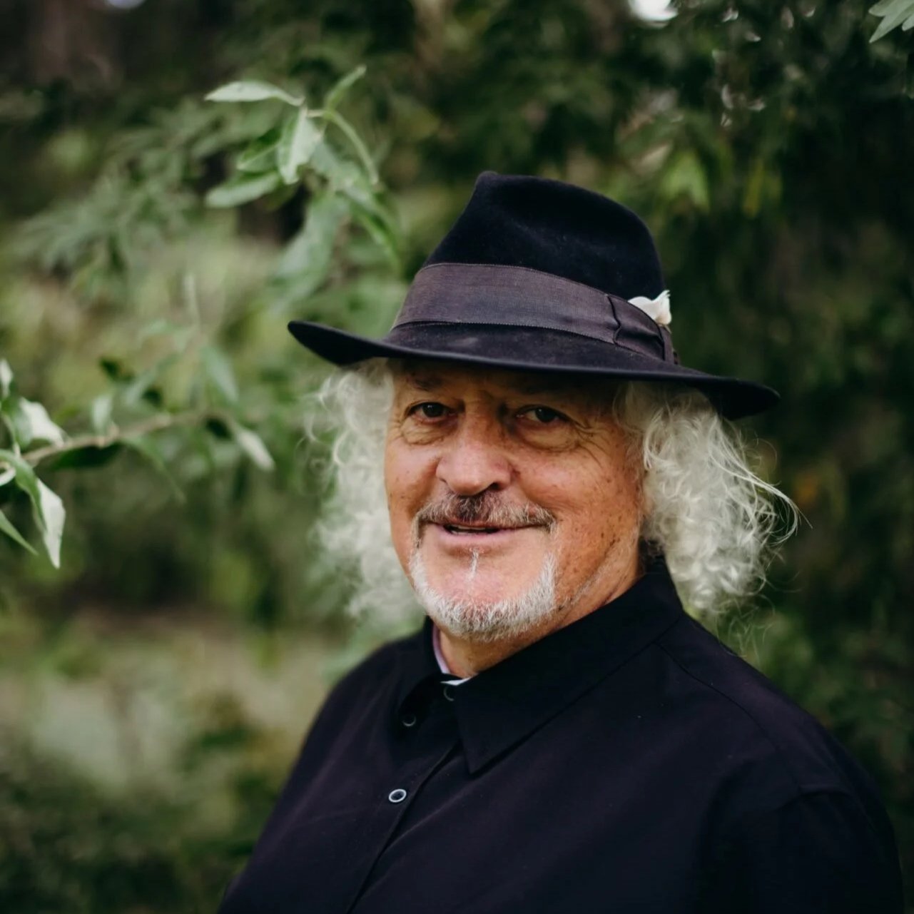 A man with curly white hair, a gray beard, and a mustache, wearing a black hat with a purple band and a black shirt, standing outdoors with greenery in the background.