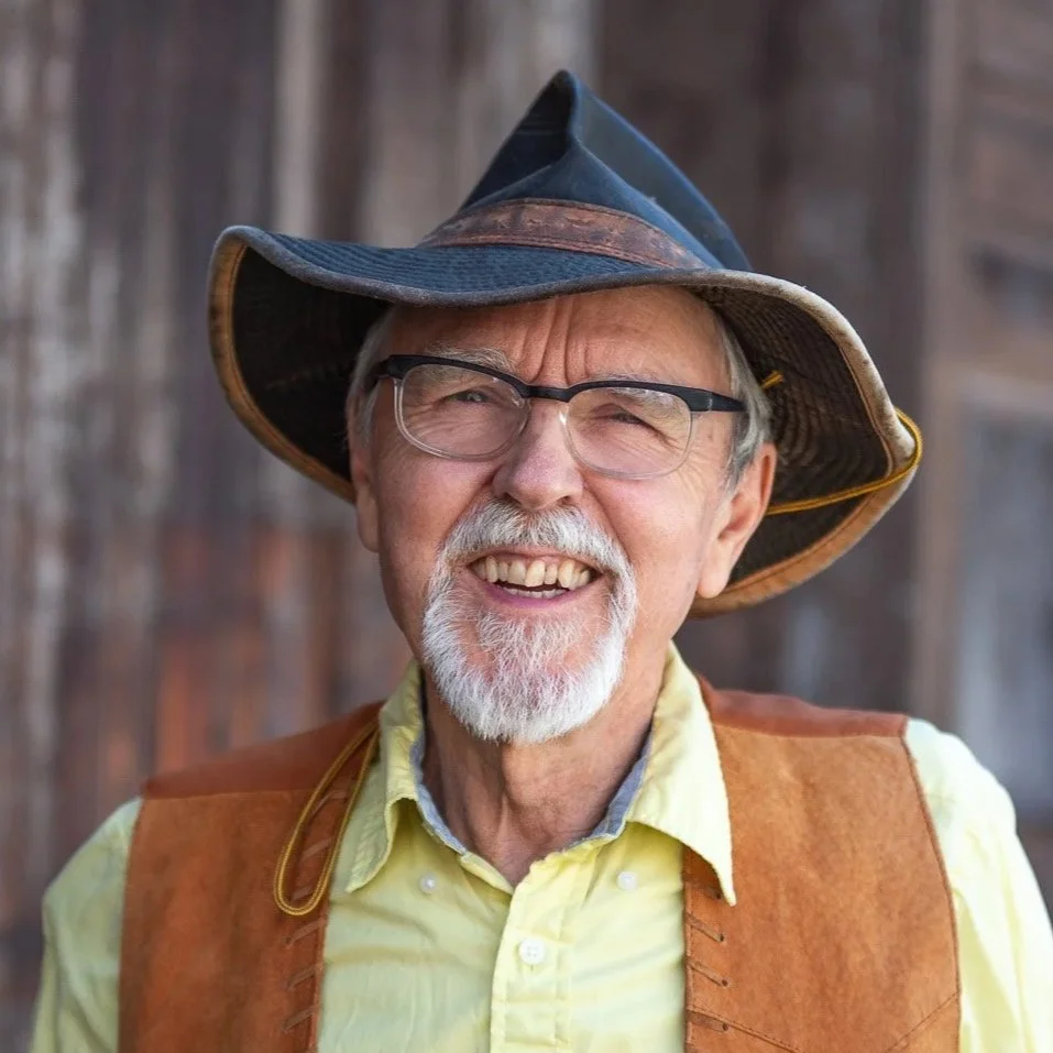 Smiling older man wearing a wide-brimmed hat, glasses, a yellow shirt, and a brown vest.