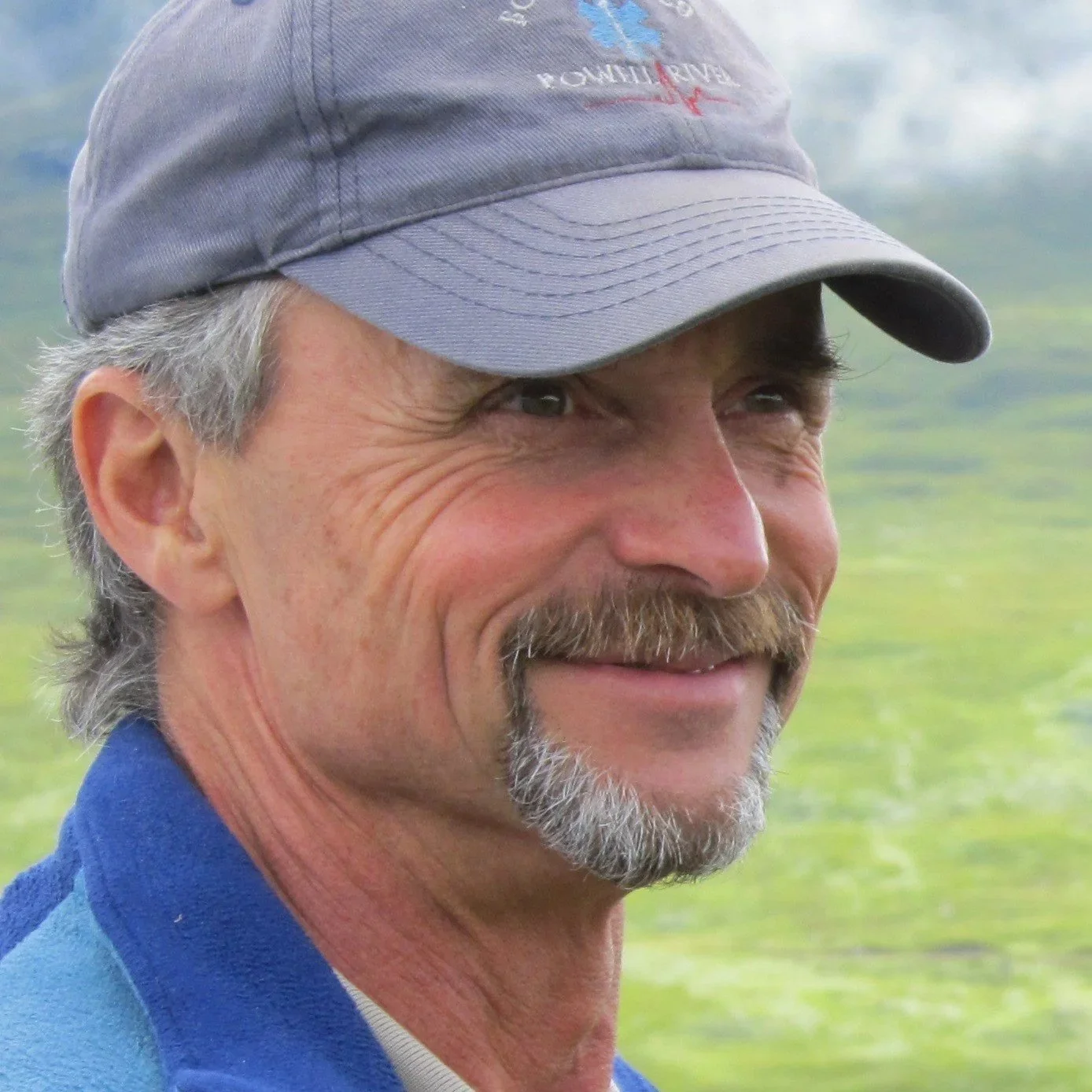 Close-up of a smiling older man with a mustache and beard, wearing a gray baseball cap and a blue jacket, outdoors in a scenic green landscape.