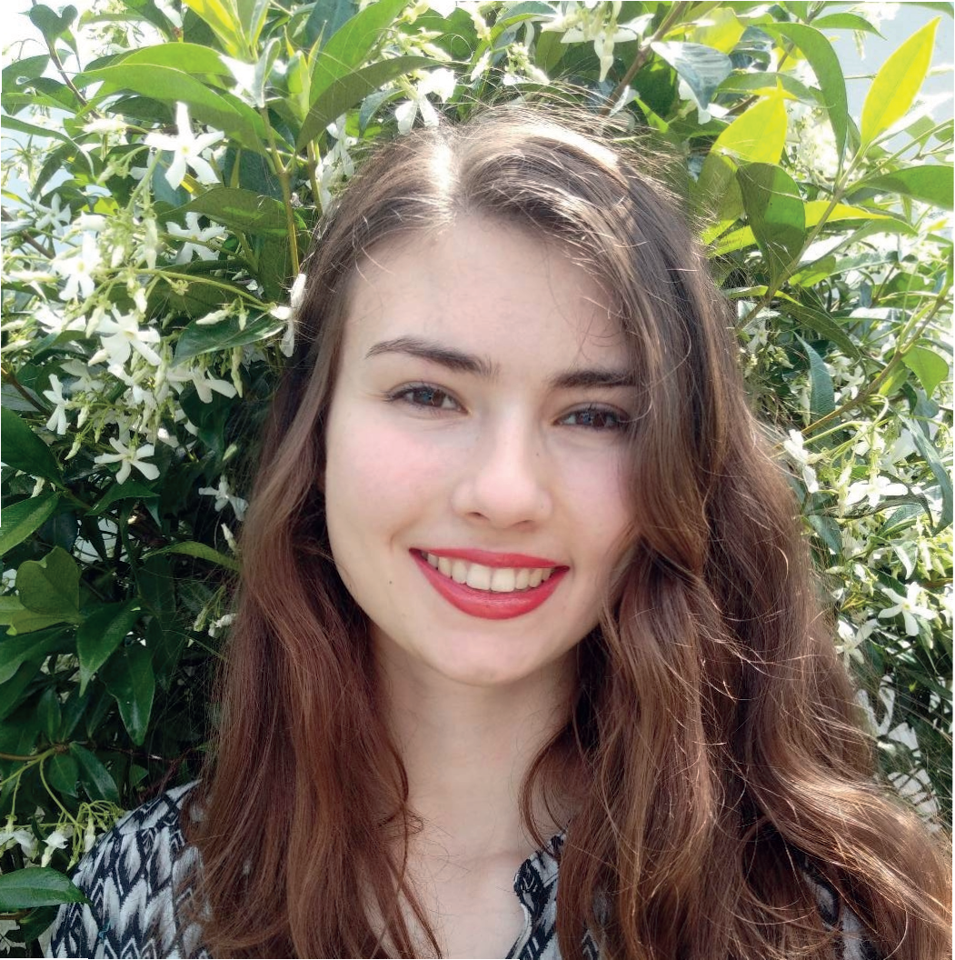 Jeune femme avec des cheveux longs et bruns, souriant, devant un buisson de feuilles vertes et de fleurs blanches.