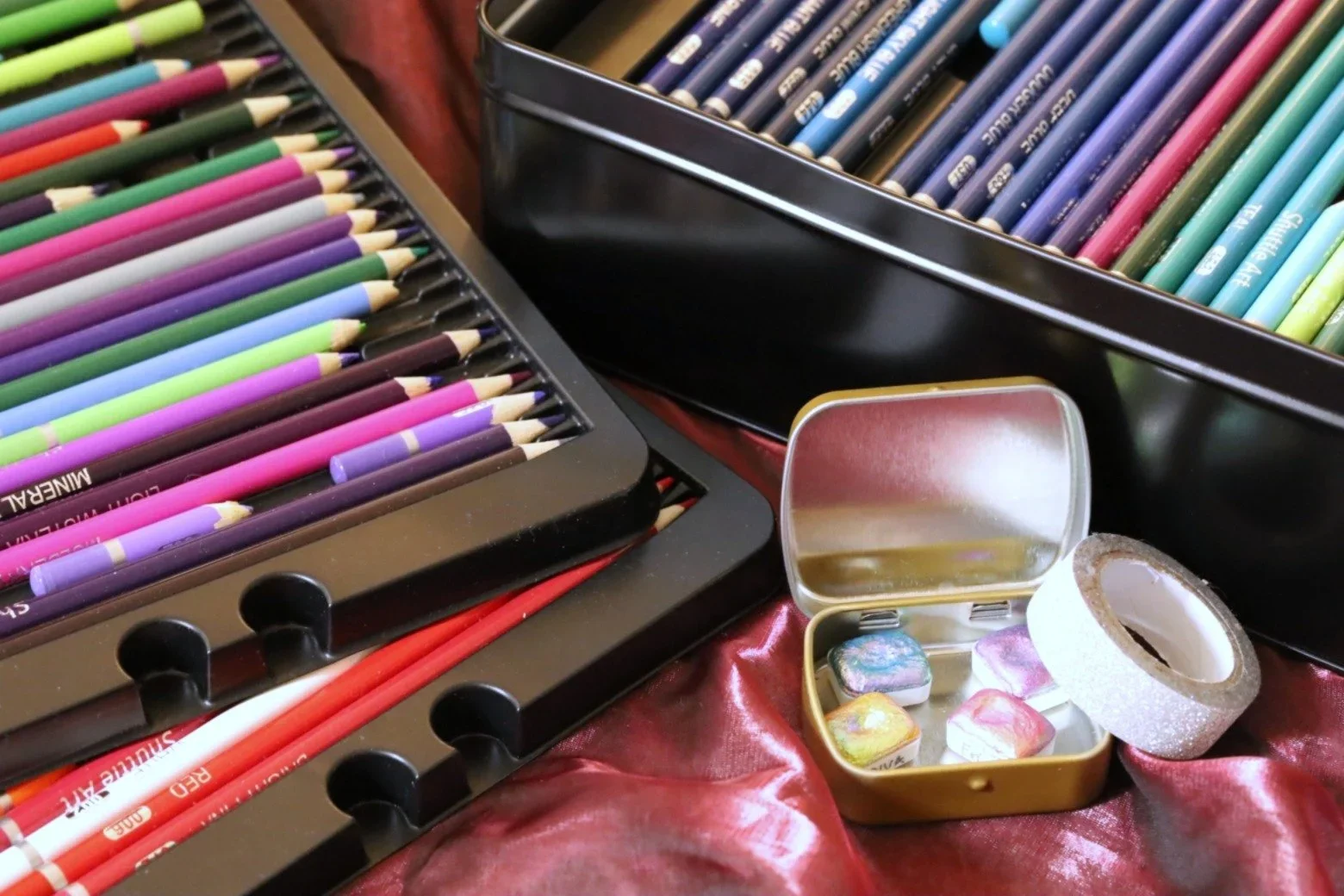 Une boîte de crayons de couleur, des godets d'aqurelle, un rouleau de ruban adhésif à paillettes, le tout disposé sur une surface en tissu rouge.