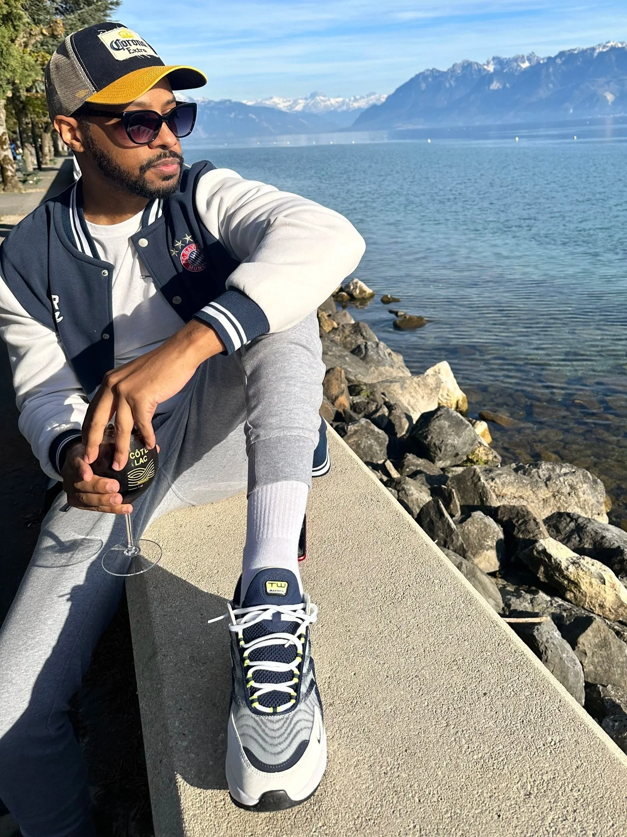Durell Arrington sitting on a concrete ledge by a lake with mountains in the background, wearing sunglasses, a baseball cap, a varsity jacket, gray sweatpants, and white sneakers, with one foot resting on the ledge.