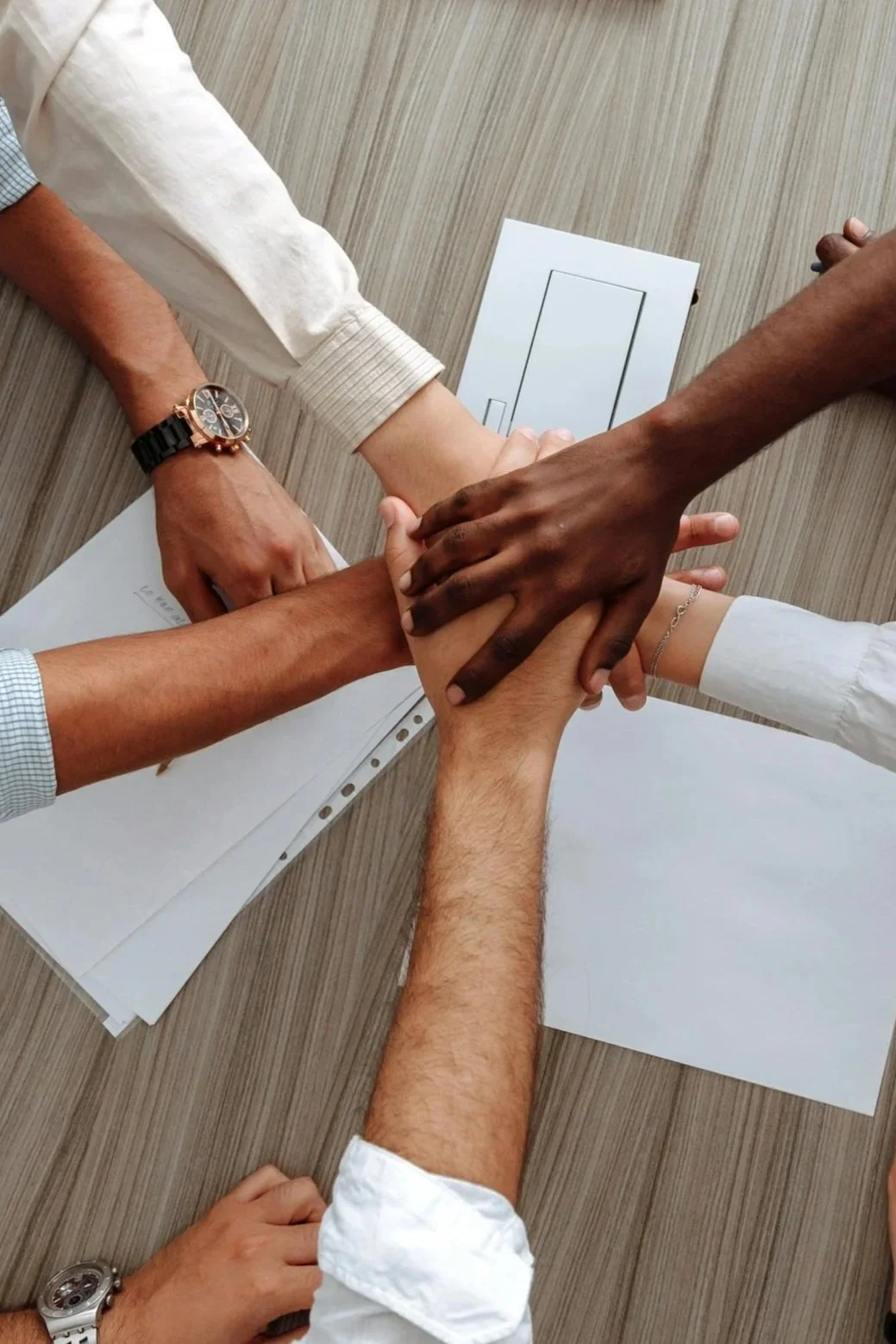 Multiple hands of diverse individuals stacked together in a show of unity, on a wooden surface with papers and a small table plug nearby. Concord NC promises12 DWI