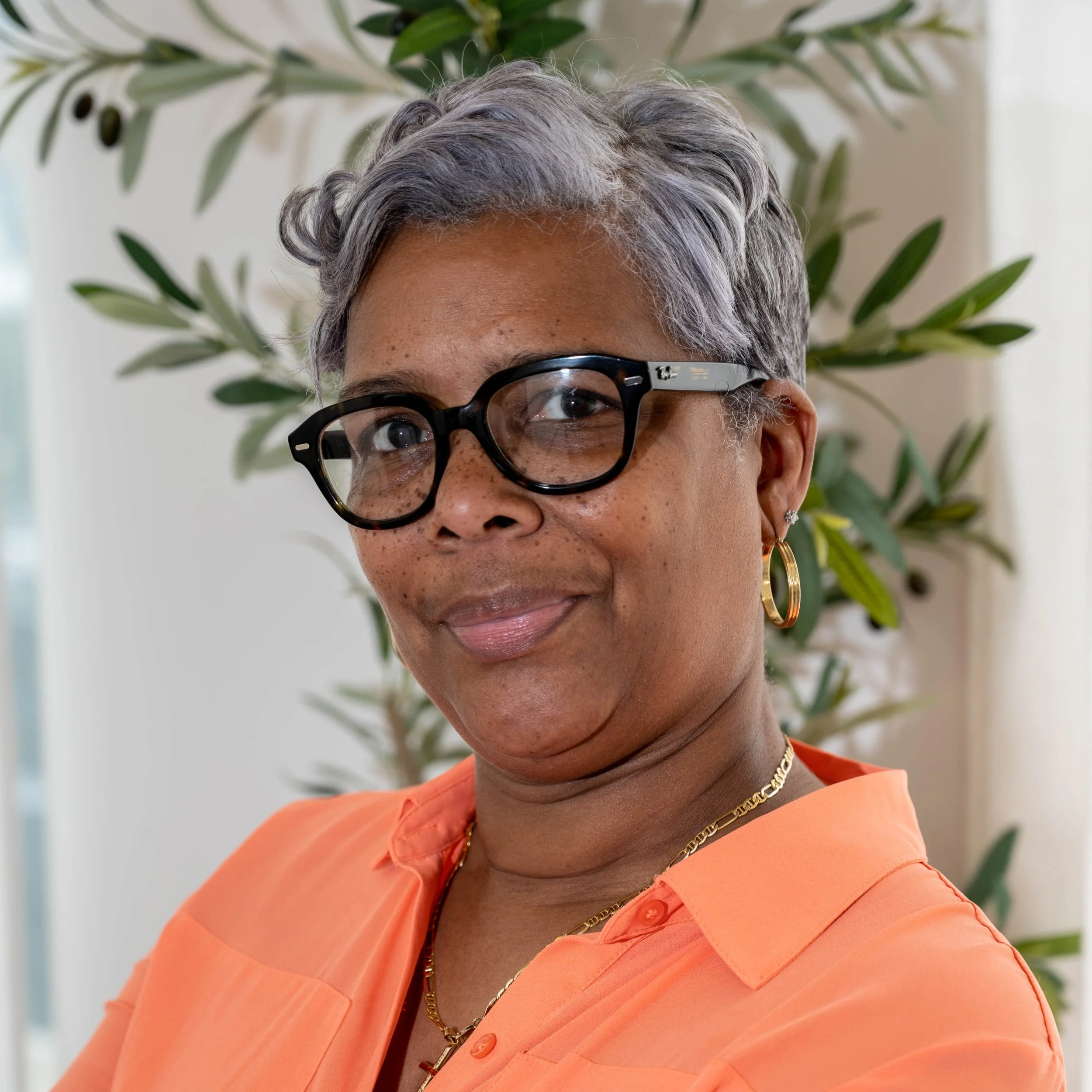 Close-up of a woman with short gray hair, wearing black glasses with tortoise shell accents, gold hoop earrings, gold chain necklace, and an orange shirt with button detail, standing in front of a leafy plant. Concord NC promises12 DWI