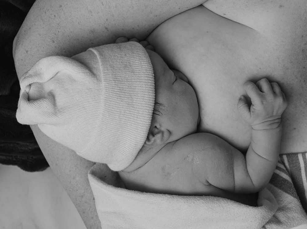 Black and white photo of a newborn baby sleeping on an adult's chest, wearing a knit cap, with a tiny fist near its face. home birth birth photographer 