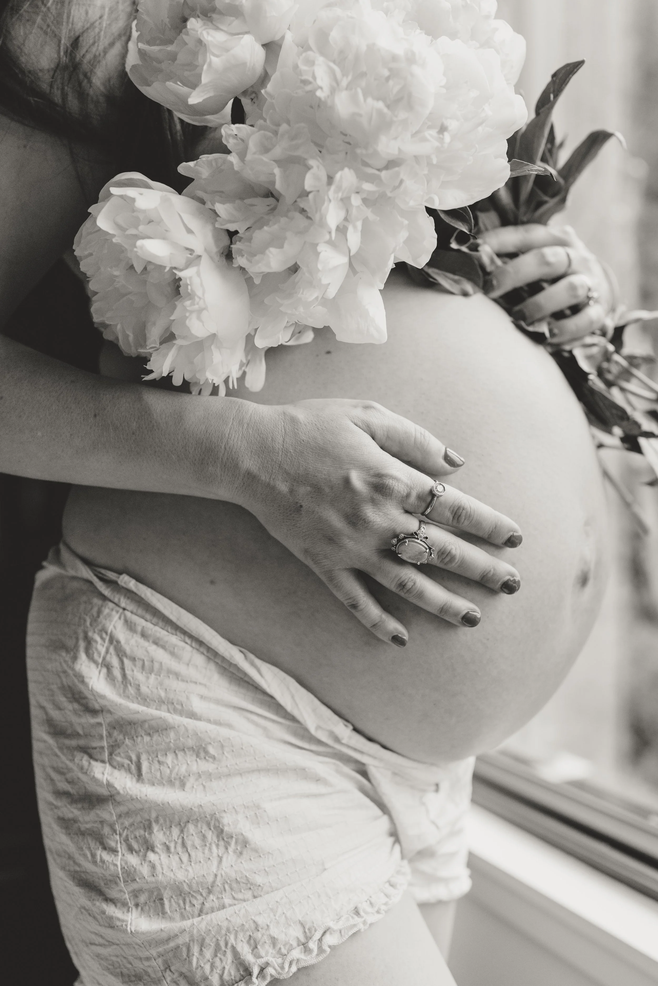 black and white photo of pregnant woman's exposed belly. she is covering her chest with flowers while cradling her belly with her other hand.