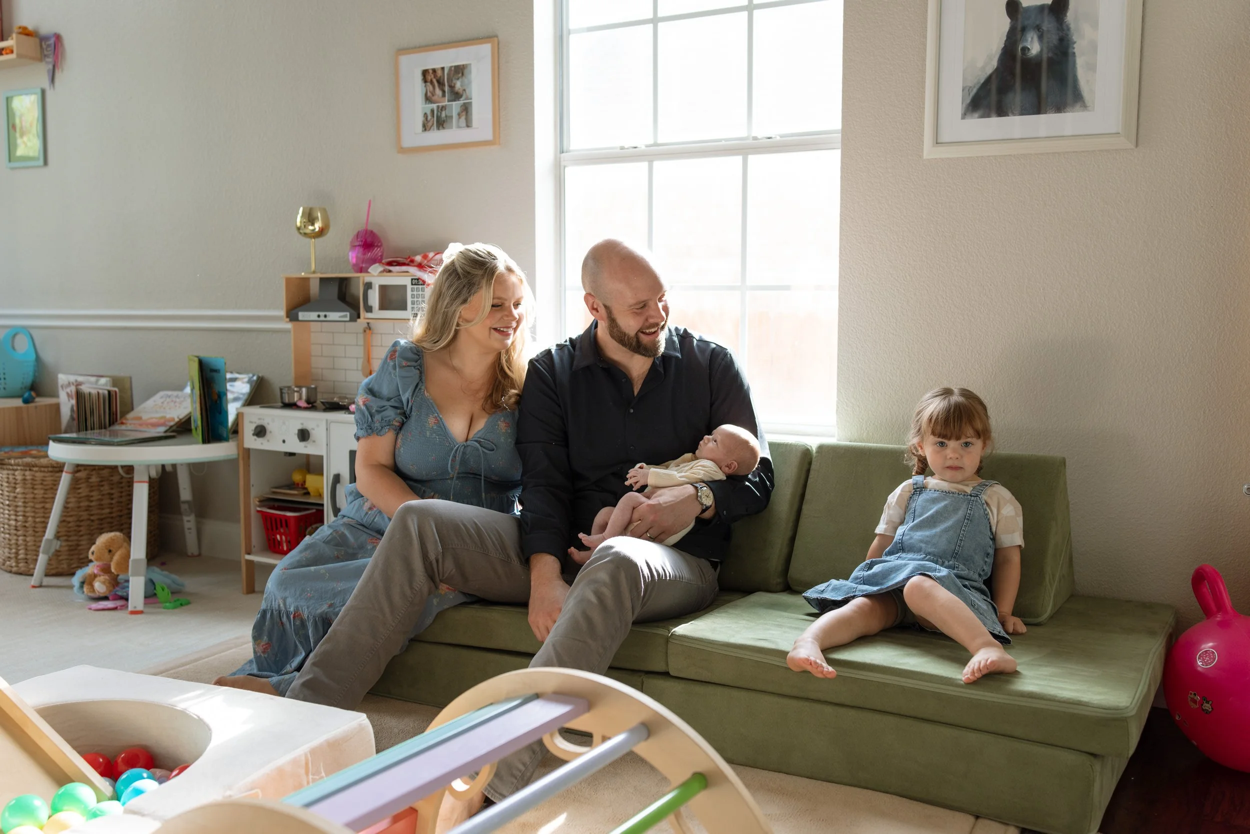 A family of four, including two young daughters, sitting on a green couch in a brightly lit living room, with the father holding a baby and both parents smiling. The children sit on the floor nearby, one looking at the camera, as toys are scattered a