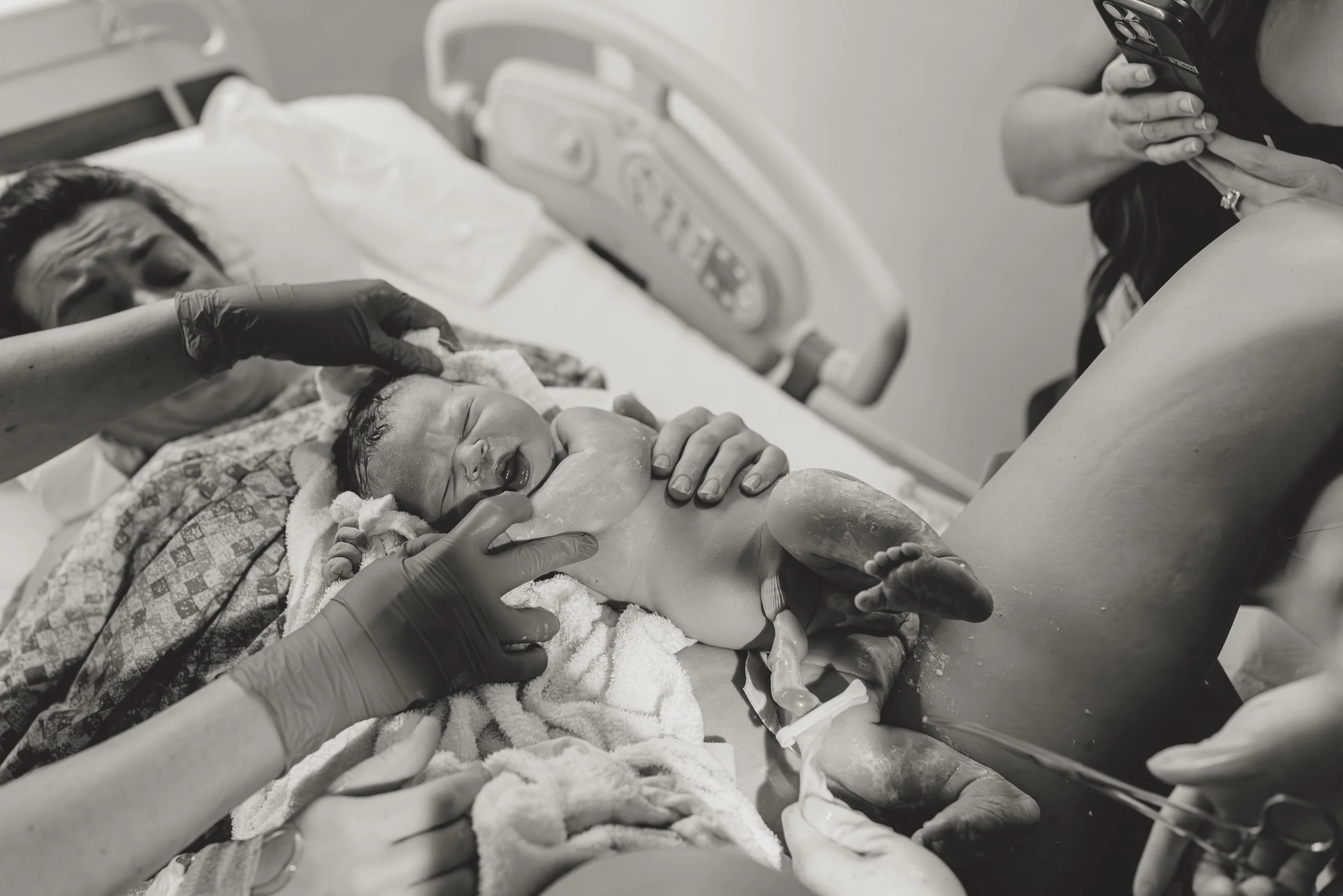 A newborn baby lying in a hospital bed with medical staff attending, surrounded by medical equipment, with a woman taking a photo.