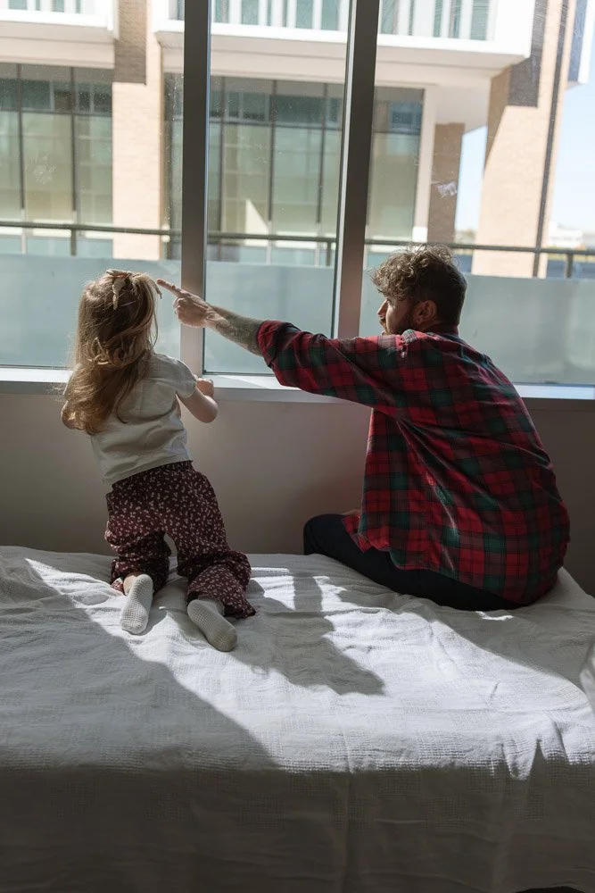 A young girl kneels on a bed, facing a large window, while a man with curly hair and a beard, wearing a red and black plaid shirt, sits next to her, reaching out touch the window. The scene is brightly lit by natural sunlight, with buildings visible 