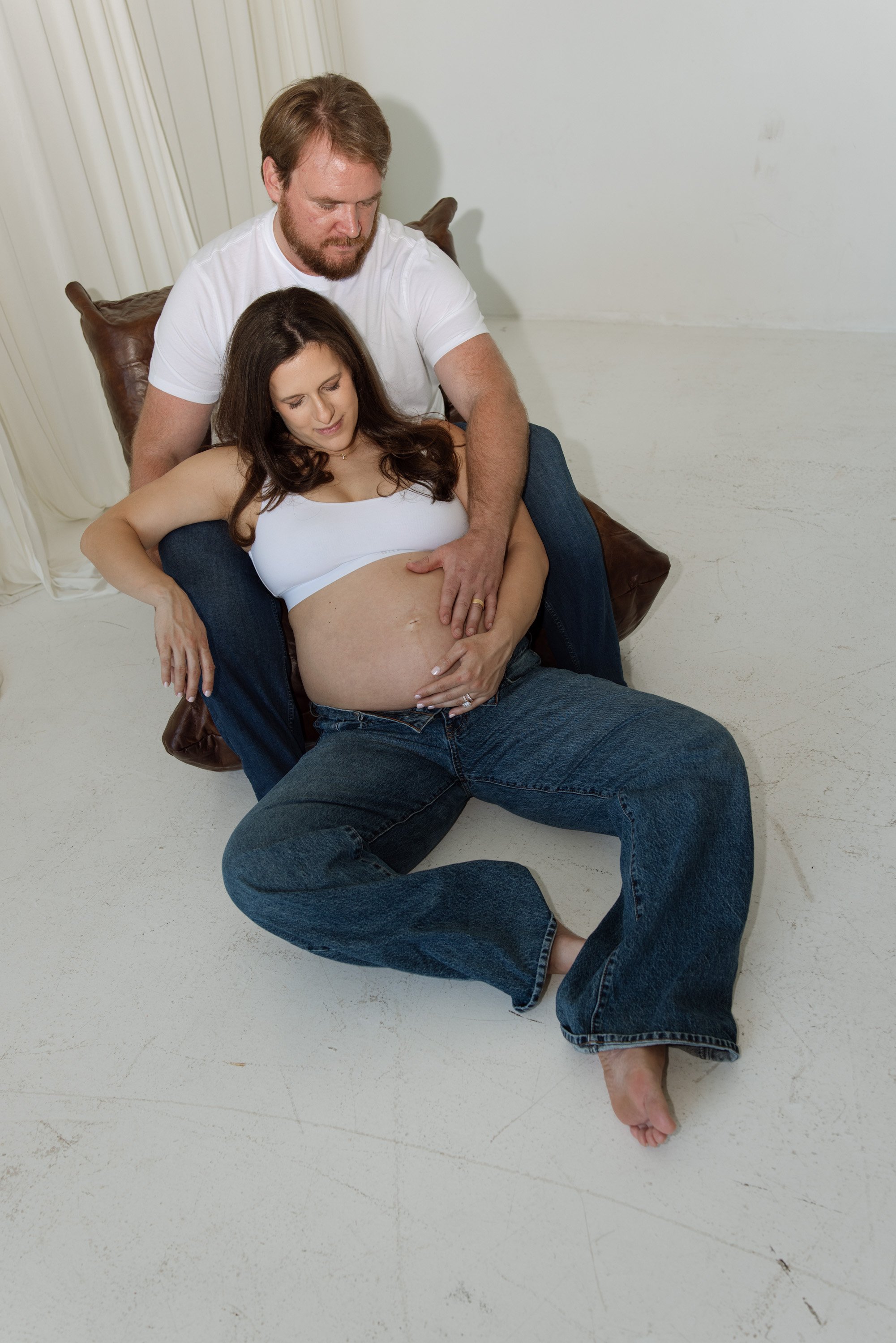 A pregnant woman lying on her back on a floor with her eyes closed, while a man is holding her belly and sitting behind her, with his hand on her abdomen and his head close to her. editorial studio maternity photography in Austin 