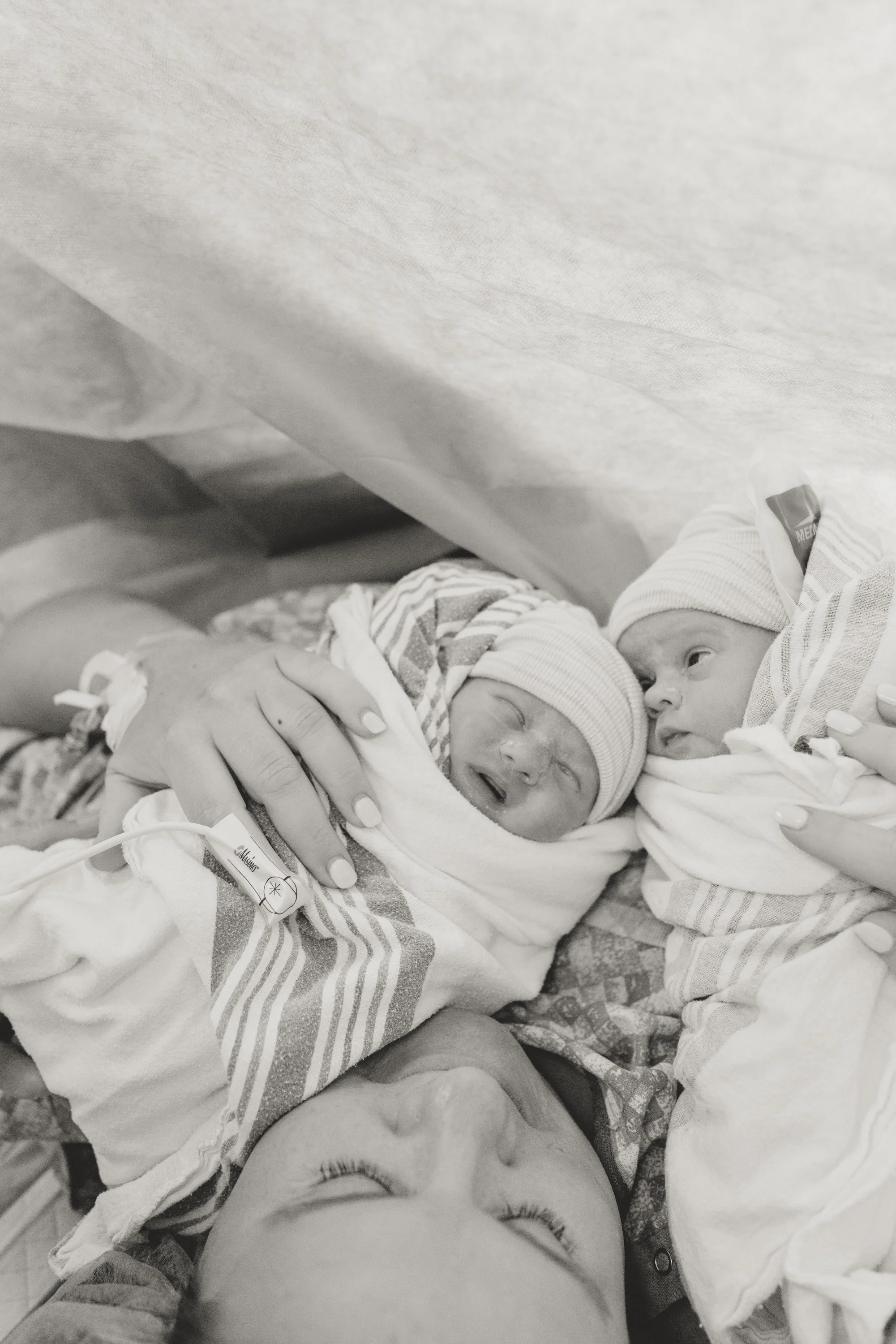 Two newborn babies in hospital bassinets with medical monitors, wrapped in blankets and wearing hospital hats, lying close to each other, one appears to be crying and the other has eyes open. dripping springs birth photography 