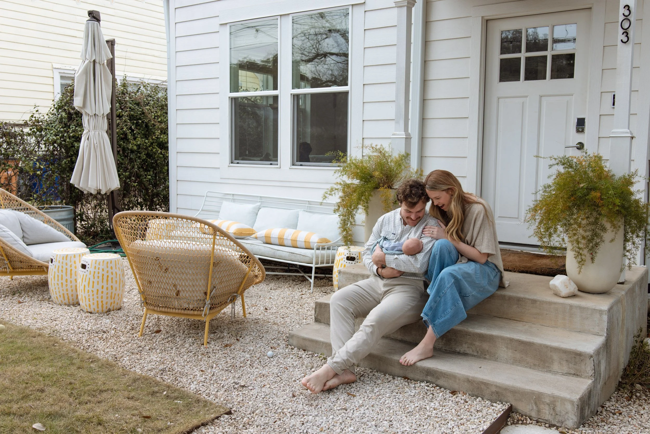 Family sitting on front steps of a house, smiling and playing with a baby. Outdoor patio with wicker furniture and yellow patterned stools, plants in large pots, and a closed umbrella. lifestyle newborn photographer in Austin 
