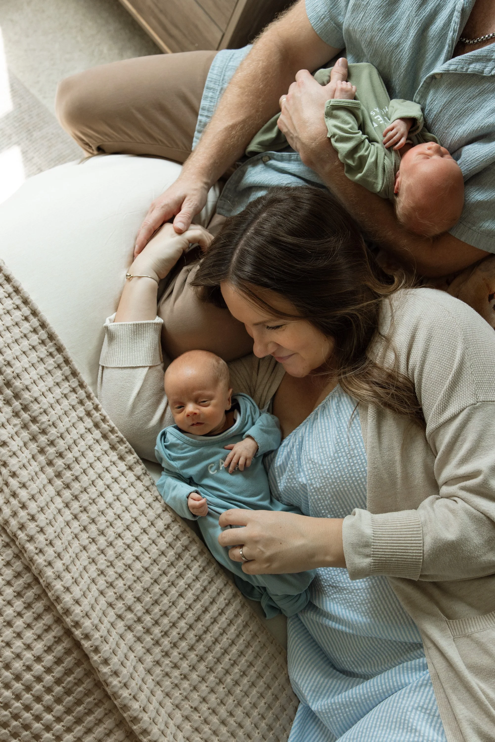 A woman and a man are lying on the floor with two babies. One baby is awake and held by the woman, and the other is sleeping on the man's chest. They are all close together, sharing a moment of affection and comfort.