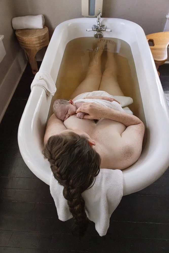 A woman lying in a vintage clawfoot bathtub filled with water, holding a newborn baby, with a towel draped over the side of the tub and a dark hardwood floor below. north Austin birth photographer 