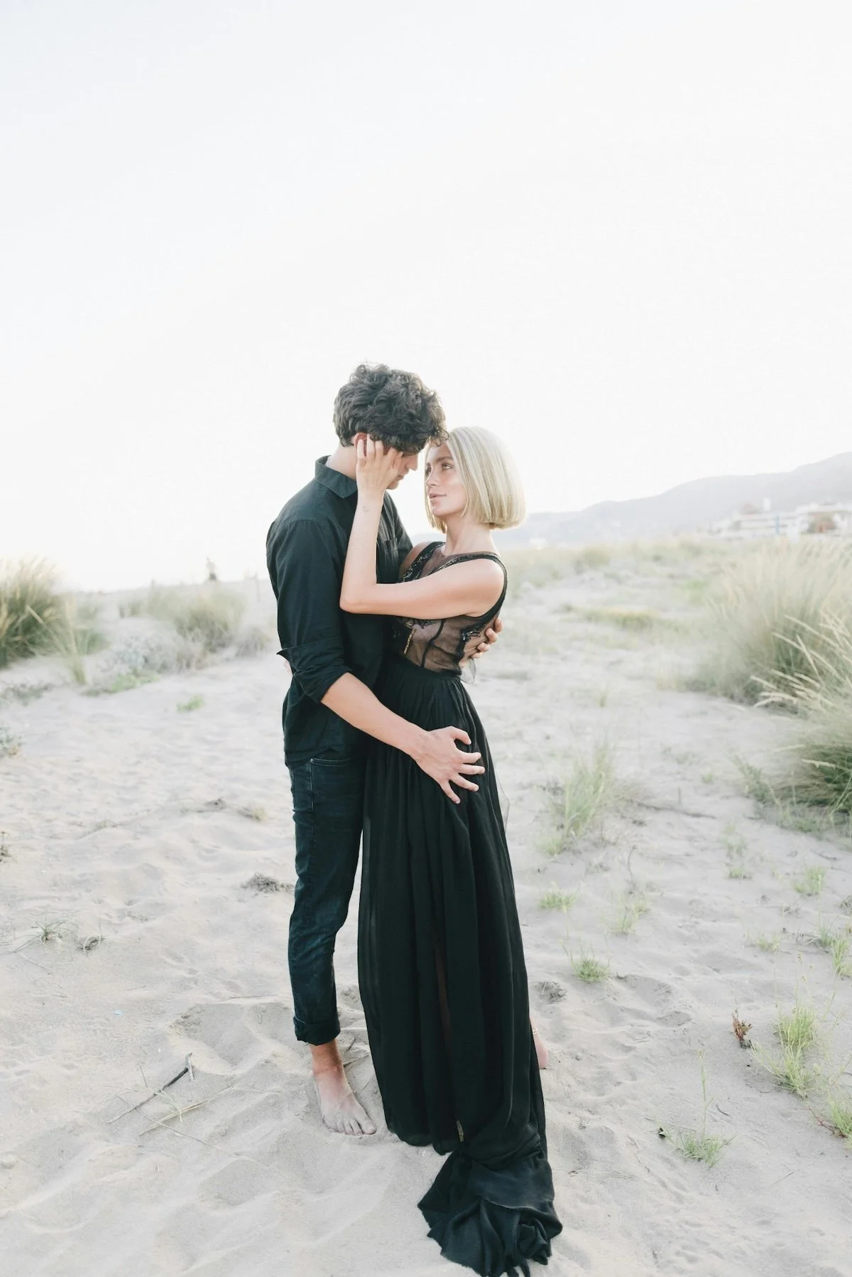 A man and woman standing close together on a sandy beach with grass and dunes in the background. The man is wearing a black shirt and pants, and the woman is wearing a black dress with lace details. They are gazing into each other's eyes, embracing l
