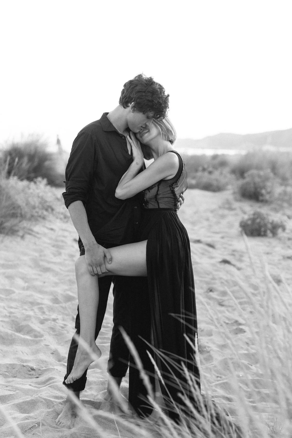 A black-and-white photo of a couple embracing passionately on a sandy beach, with hills in the background.