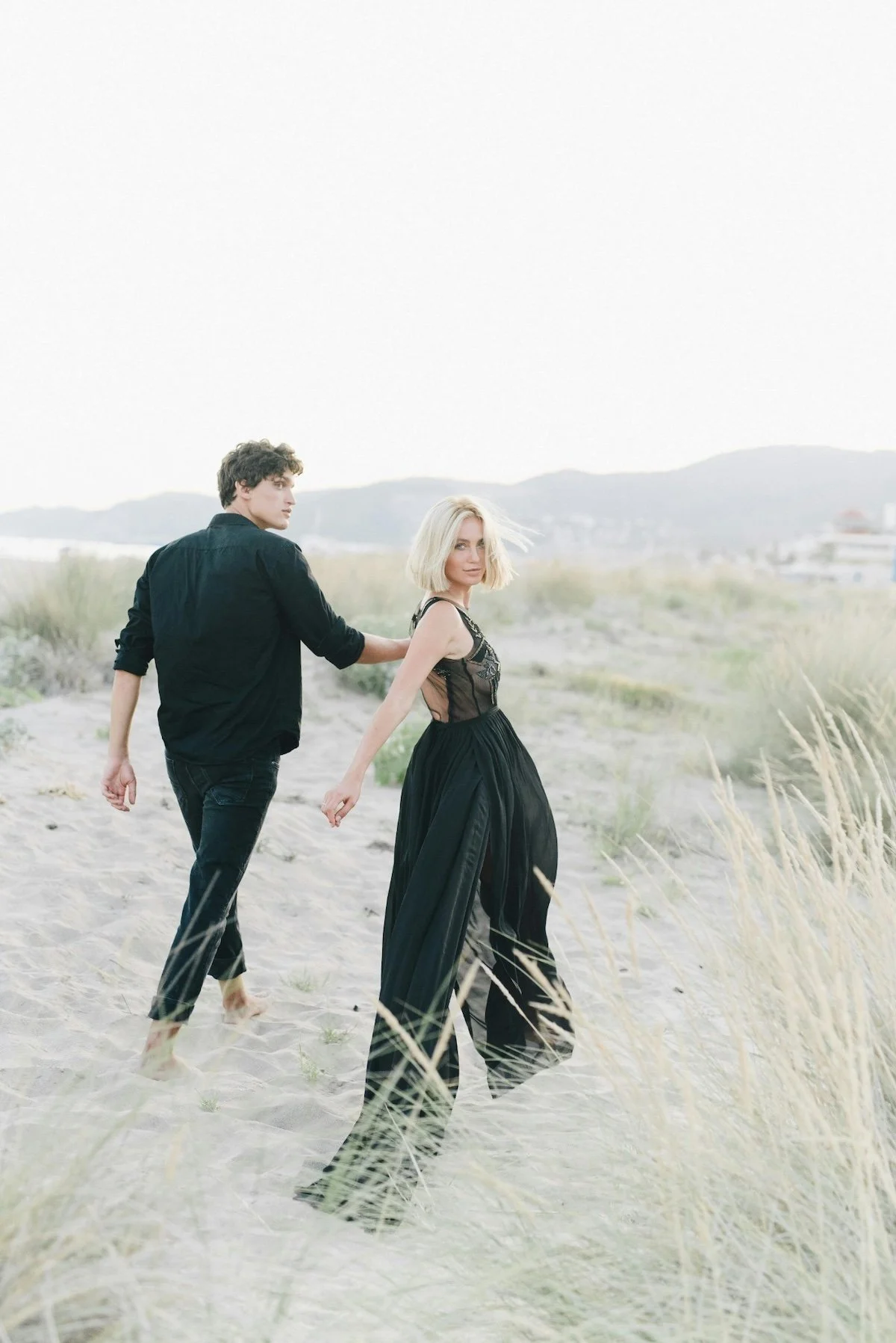 A man and woman with light skin holding hands on a sandy beach with grasses and distant mountains in the background. The woman wears a black dress, and the man wears black clothing.