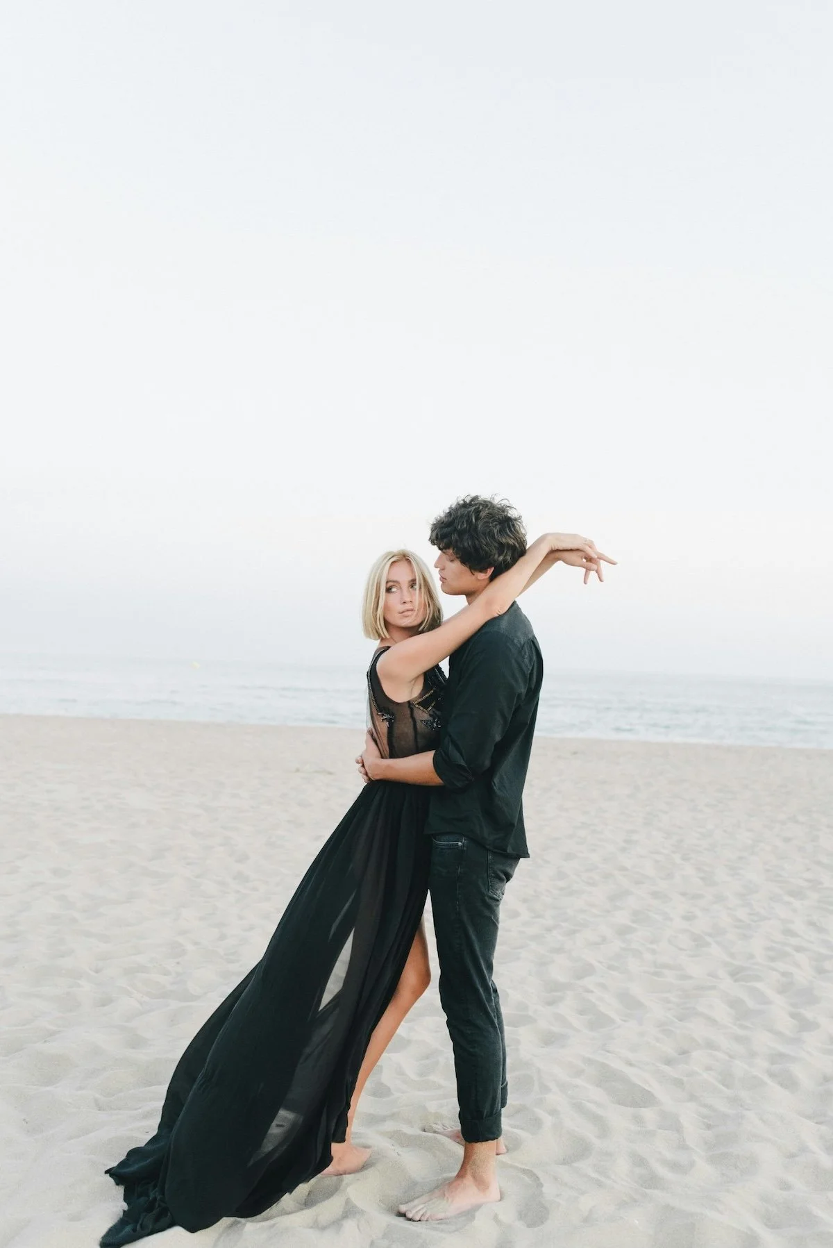 A couple stands on the beach embracing, the woman in a black dress with sheer details and the man in black shirt and pants, with the ocean and sky in the background.