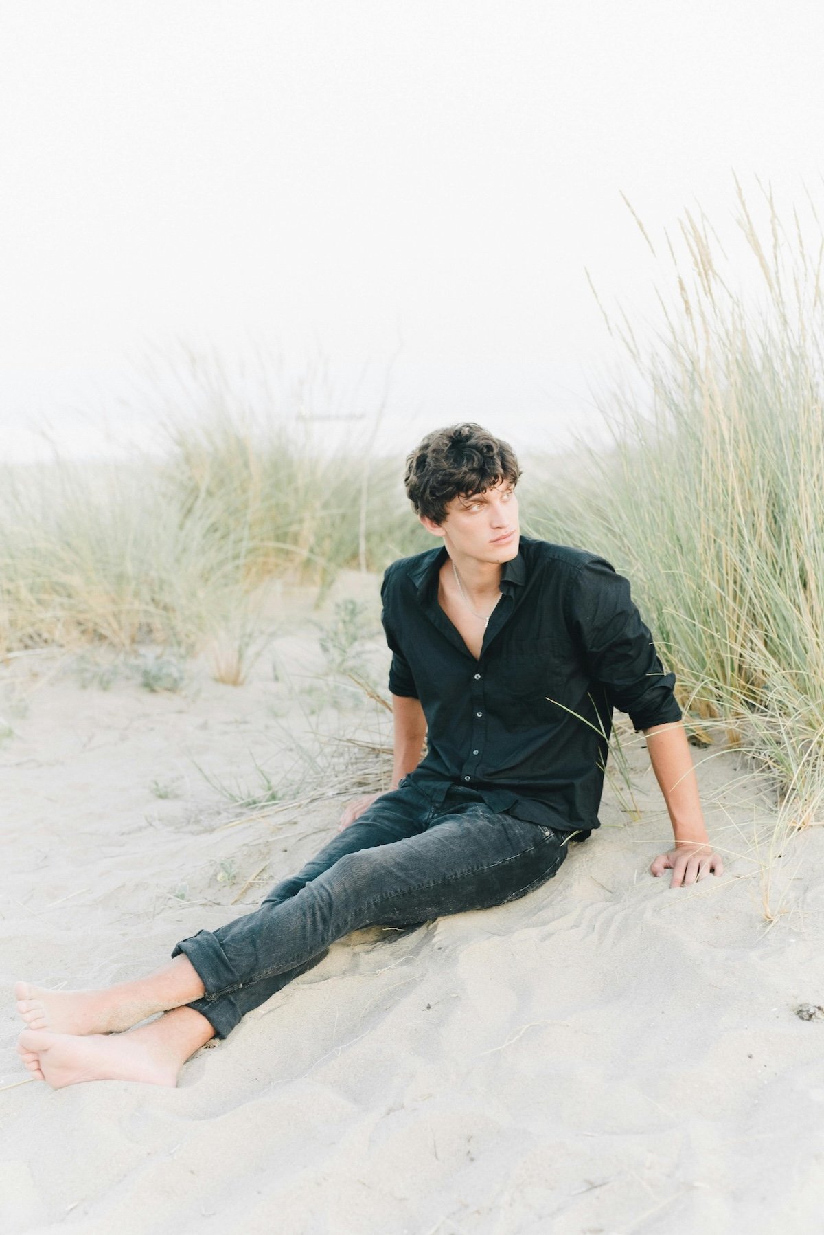 Young man with curly hair in a black shirt and jeans sitting on sandy beach amid tall grass, looking to the side.