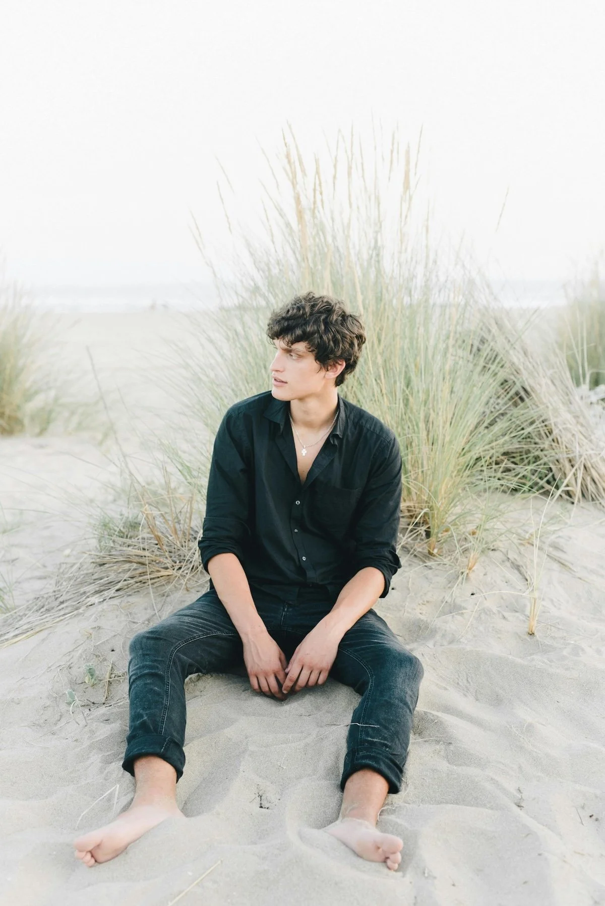 A young man with dark curly hair, wearing a black shirt and black jeans, sitting on sandy ground surrounded by tall beach grass, looking to his left.
