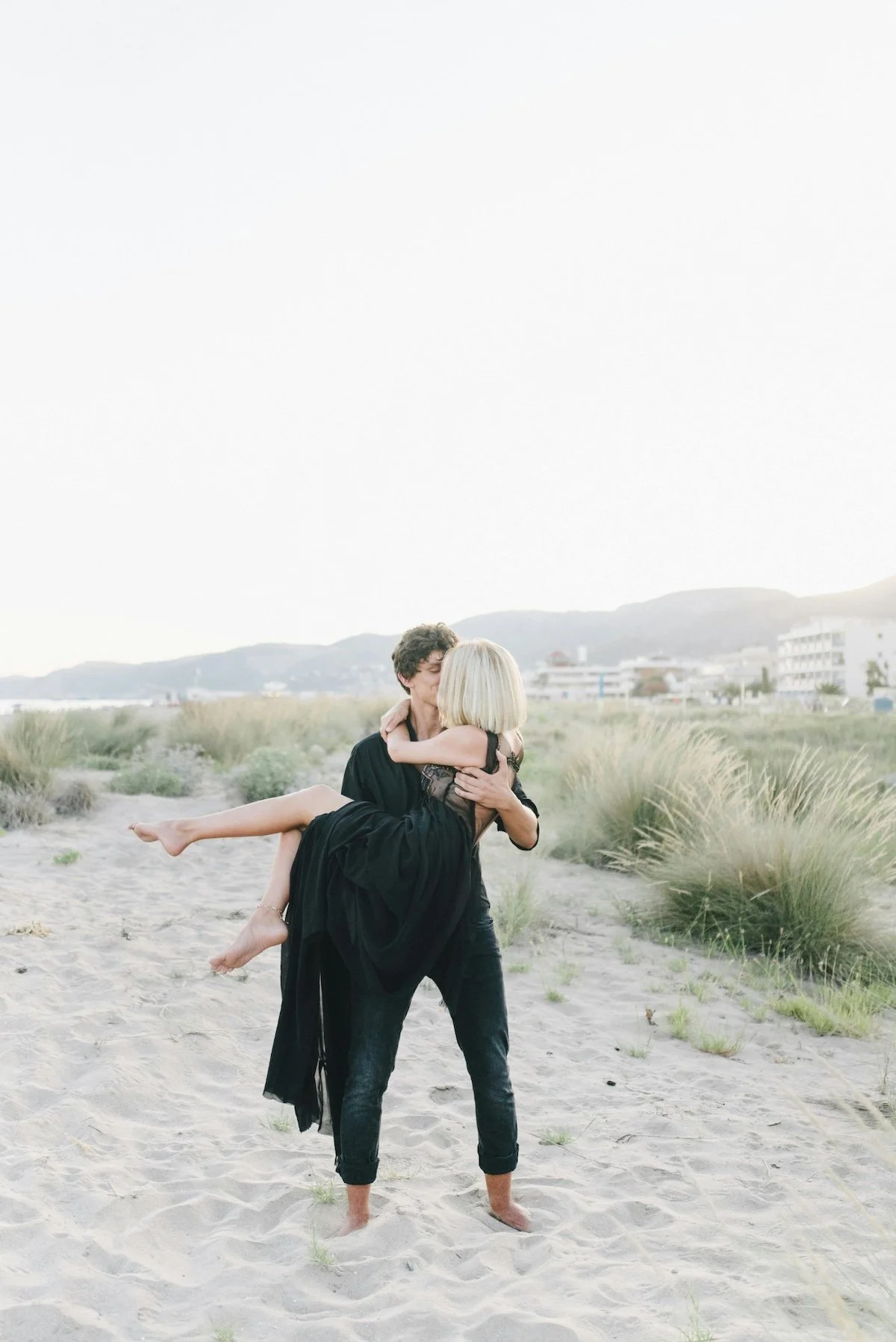 A couple on a beach, with the man lifting and holding the woman in his arms as they kiss, surrounded by sand and grassy dunes, with buildings and hills in the background.