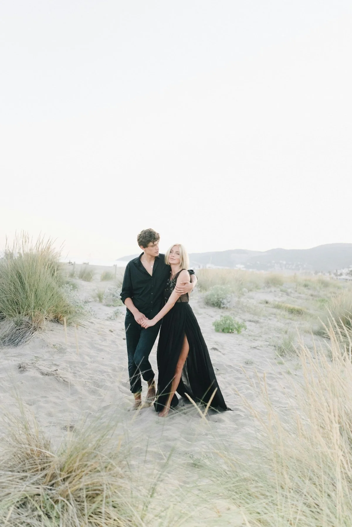 A young man and woman standing on a sandy beach with tall grasses, holding hands, the woman wearing a long black dress and the man in dark clothing, with mountains visible in the background during sunset.