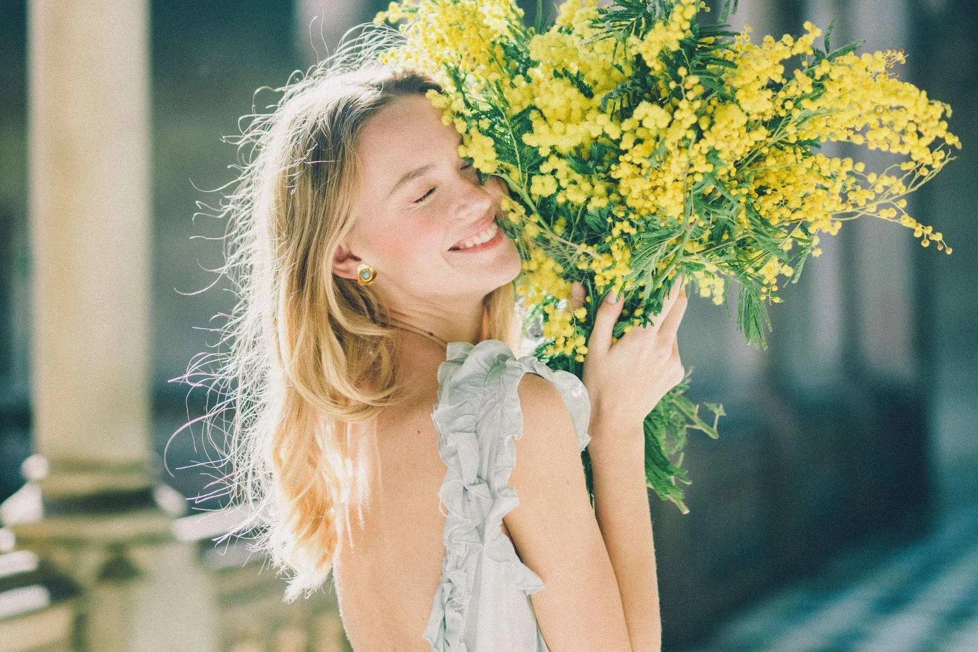 A young woman with blonde hair smiling and holding a large bouquet of yellow flowers close to her face.
